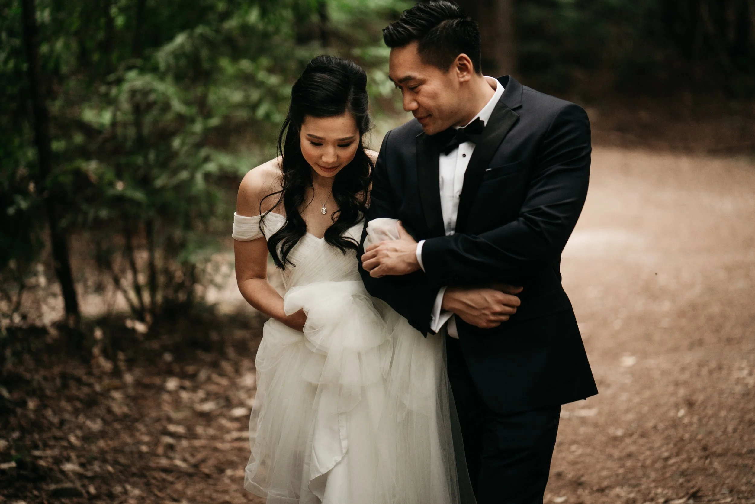 A bride and groom standing close together on a wooded path; the bride is in a white wedding dress and the groom in a black tuxedo, both smiling softly.