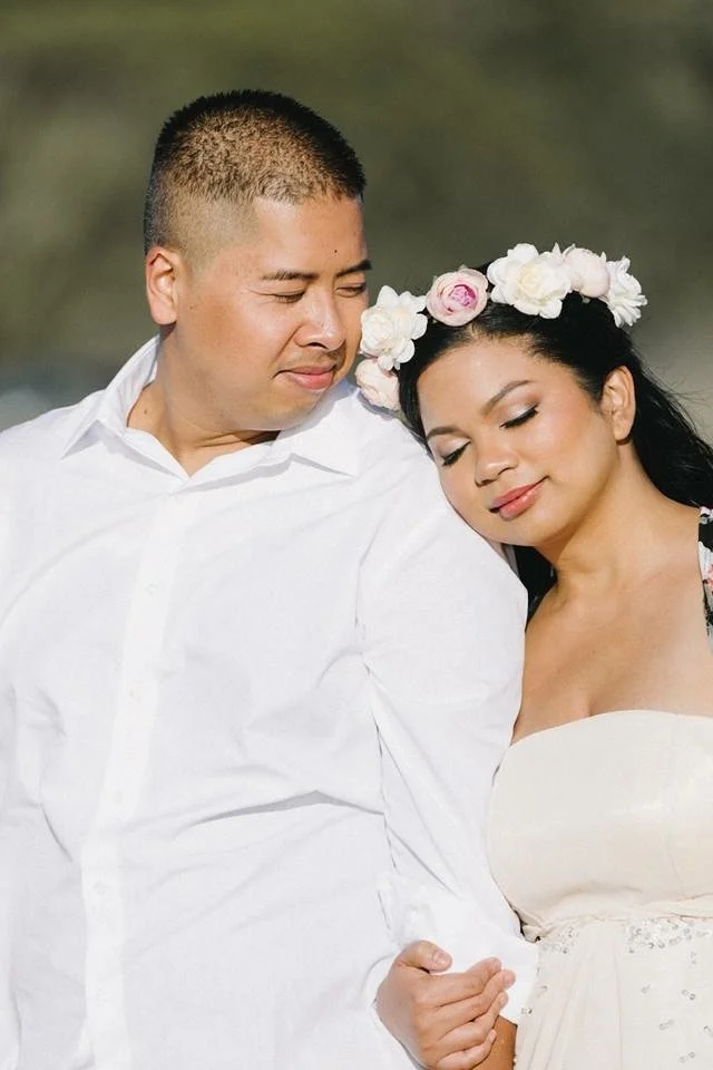 A man and woman sit close together outdoors with eyes closed, the man wearing a white shirt and the woman with a floral crown, both appearing peaceful and content.