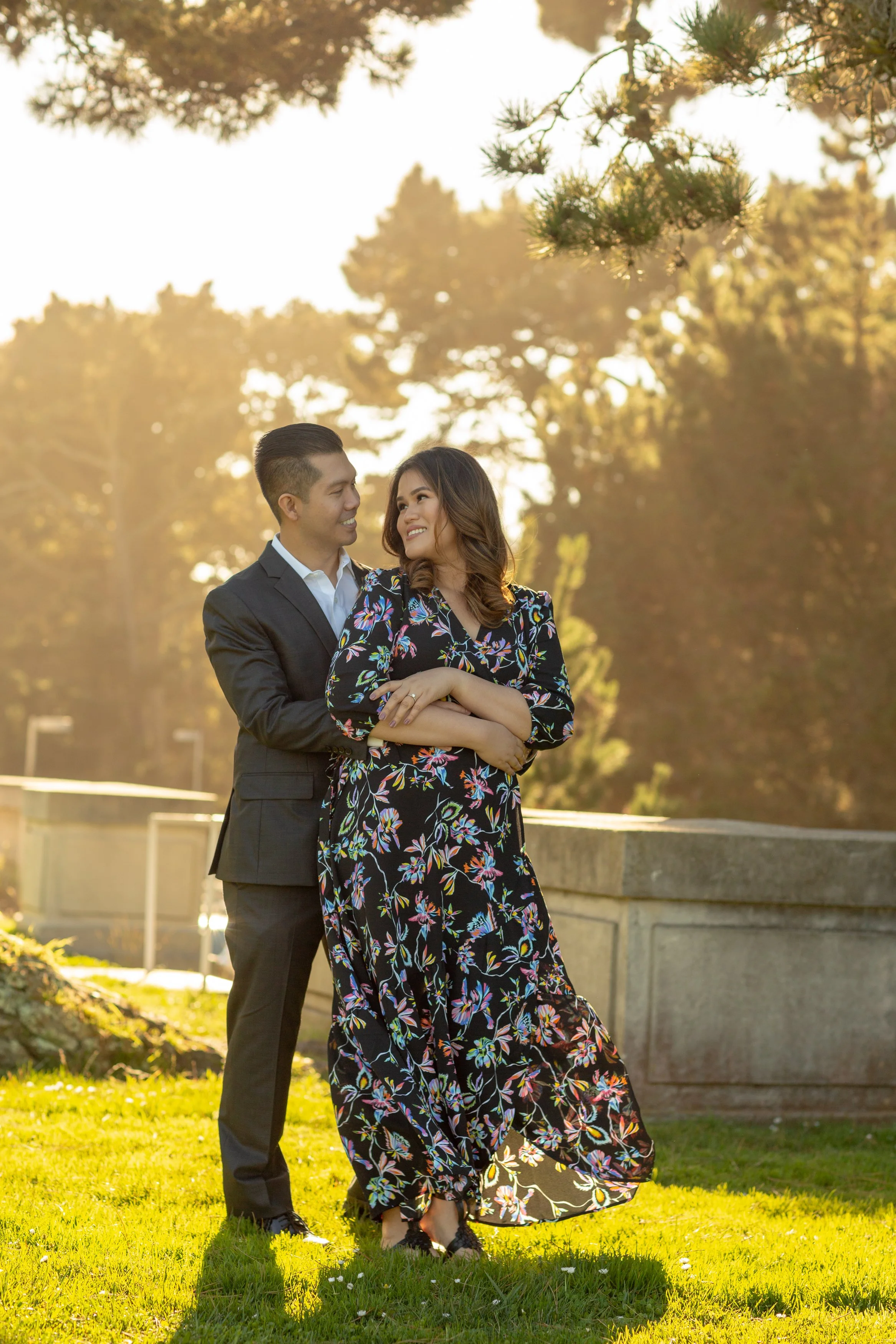 A man in a dark suit and a woman in a long, floral dress embrace outdoors during golden hour, with trees in the background.