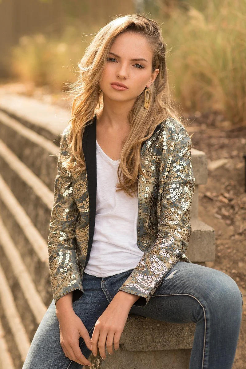 A young woman with wavy blonde hair sitting on a concrete ledge outdoors, wearing a shiny, sequined jacket, white t-shirt, and blue jeans, with a neutral expression and slightly squinted eyes.