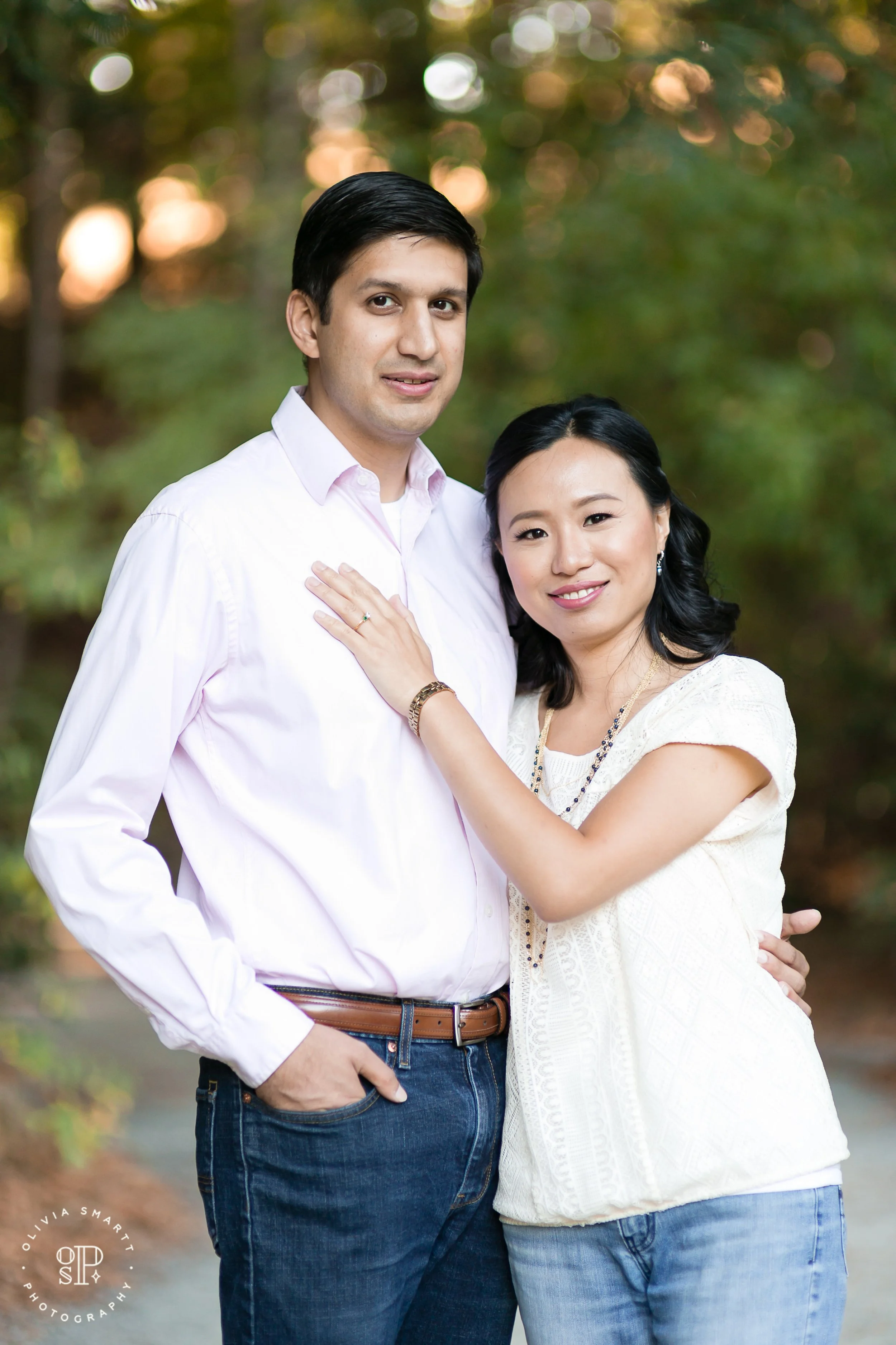 A smiling couple standing outdoors in a wooded area, with the woman resting her hand on the man's chest, both dressed casually.