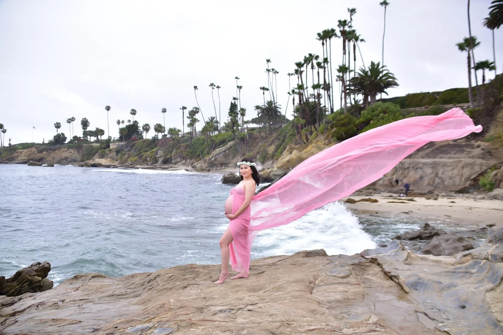 Pregnant woman in pink dress on rocky beach with pink fabric flowing in the wind, palm trees and cloudy sky in the background.