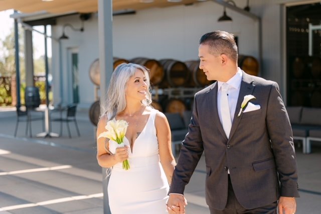 A bride and groom holding hands and smiling at each other outside a building, with wedding attire and flowers.