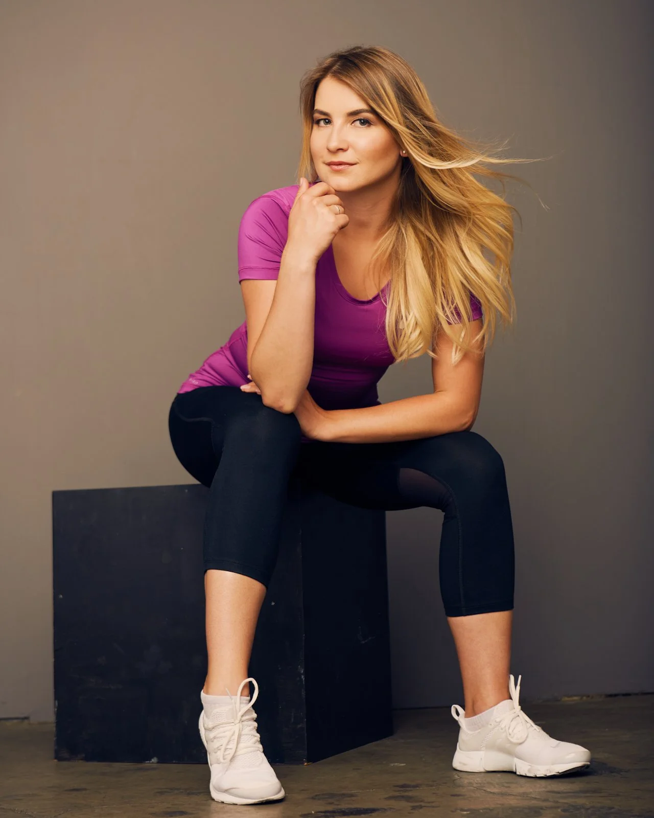 A young woman with blonde hair in a pink athletic top, black leggings, and white sneakers sitting on a black box, looking at the camera with a slight smile, in front of a plain background.