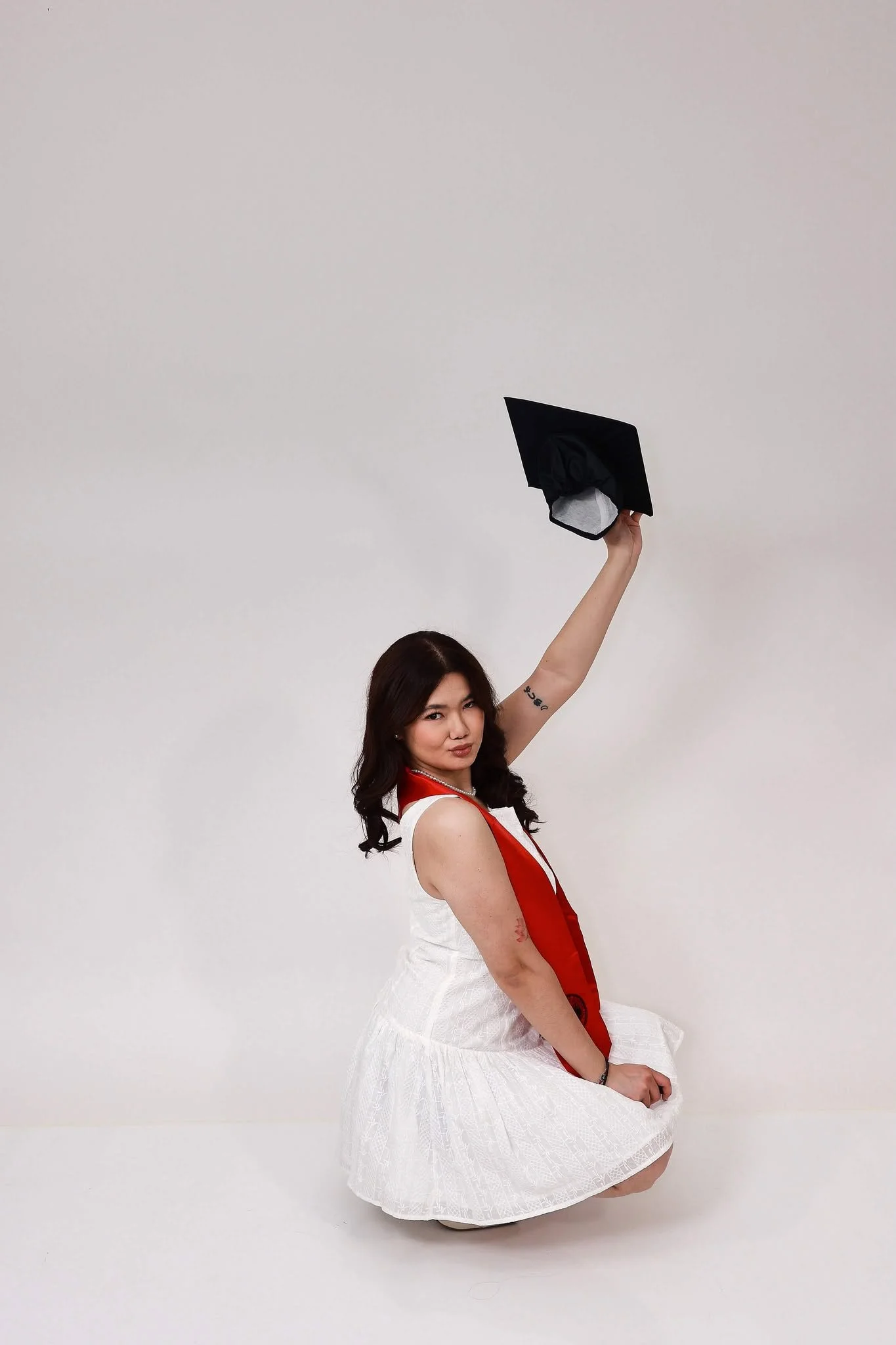 A woman wearing a white dress with a red sash, sitting on the floor, with a person behind her holding a graduation cap above their head.