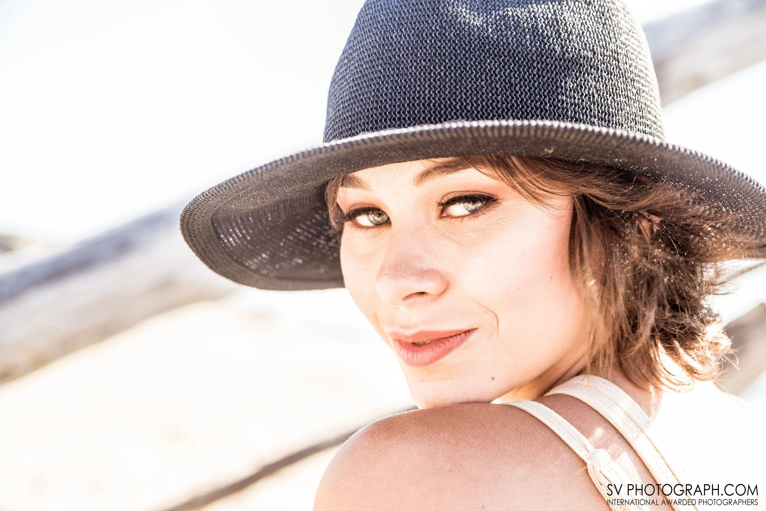 Close-up of a woman wearing a dark sun hat, smiling, with a blurred beach background.