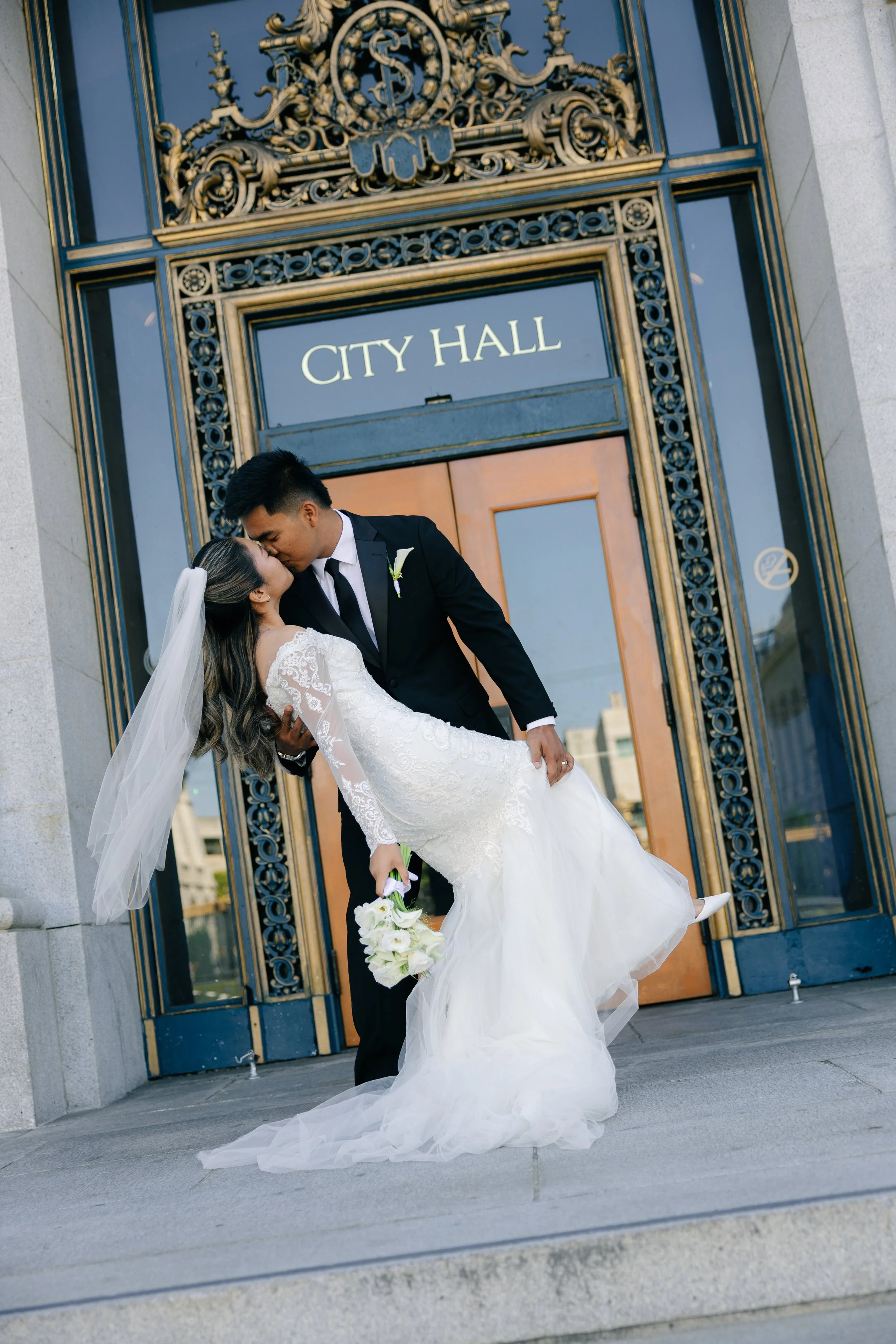A newlywed couple kissing in front of a city hall building. The groom is holding the bride in his arms, and she is holding a bouquet of white flowers. The bride is wearing a white wedding dress and veil, and the groom is in a black suit.