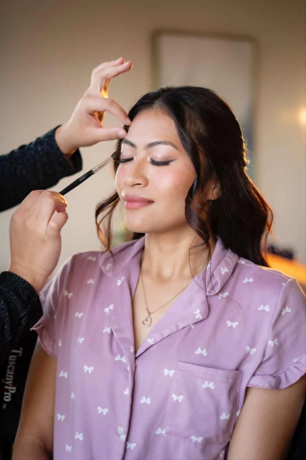A woman getting her makeup done, with her eyes closed, wearing a light purple blouse with white bow patterns and a gold necklace.