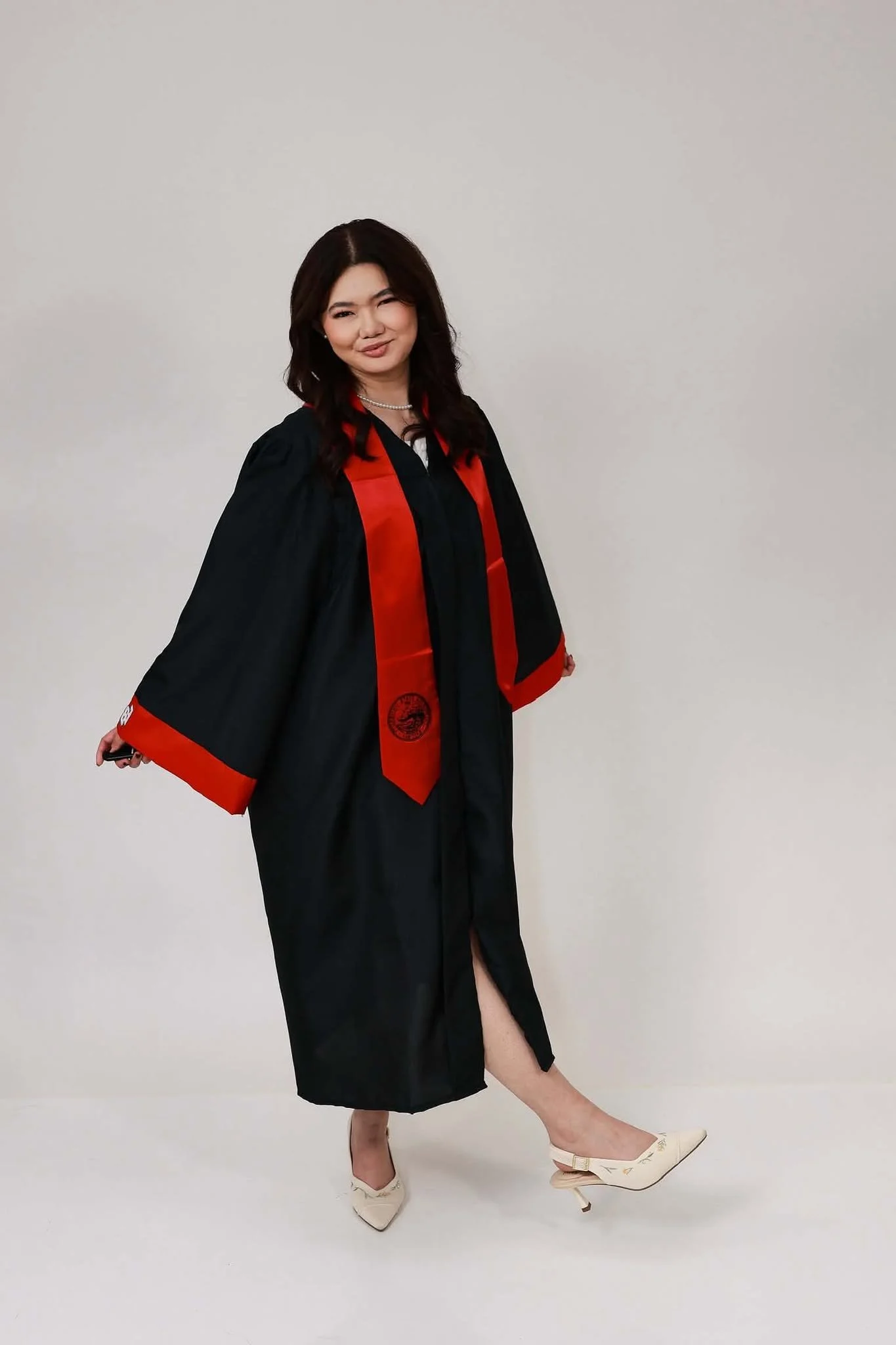 A woman in a graduation gown and cap, standing with one leg raised and wearing white heels with embroidered flowers, against a plain light-colored background.