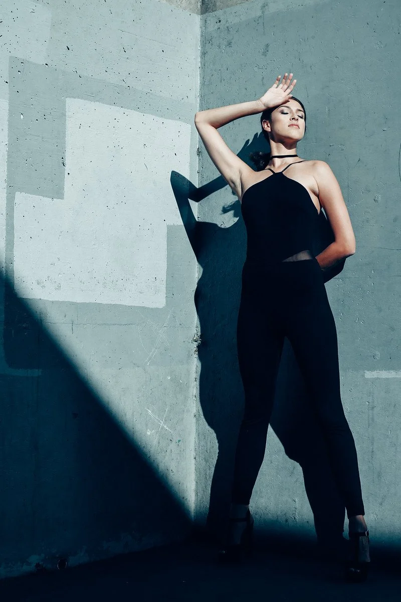A woman wearing a black jumpsuit posing against a concrete wall with one arm raised to her forehead, casting a shadow on the wall.