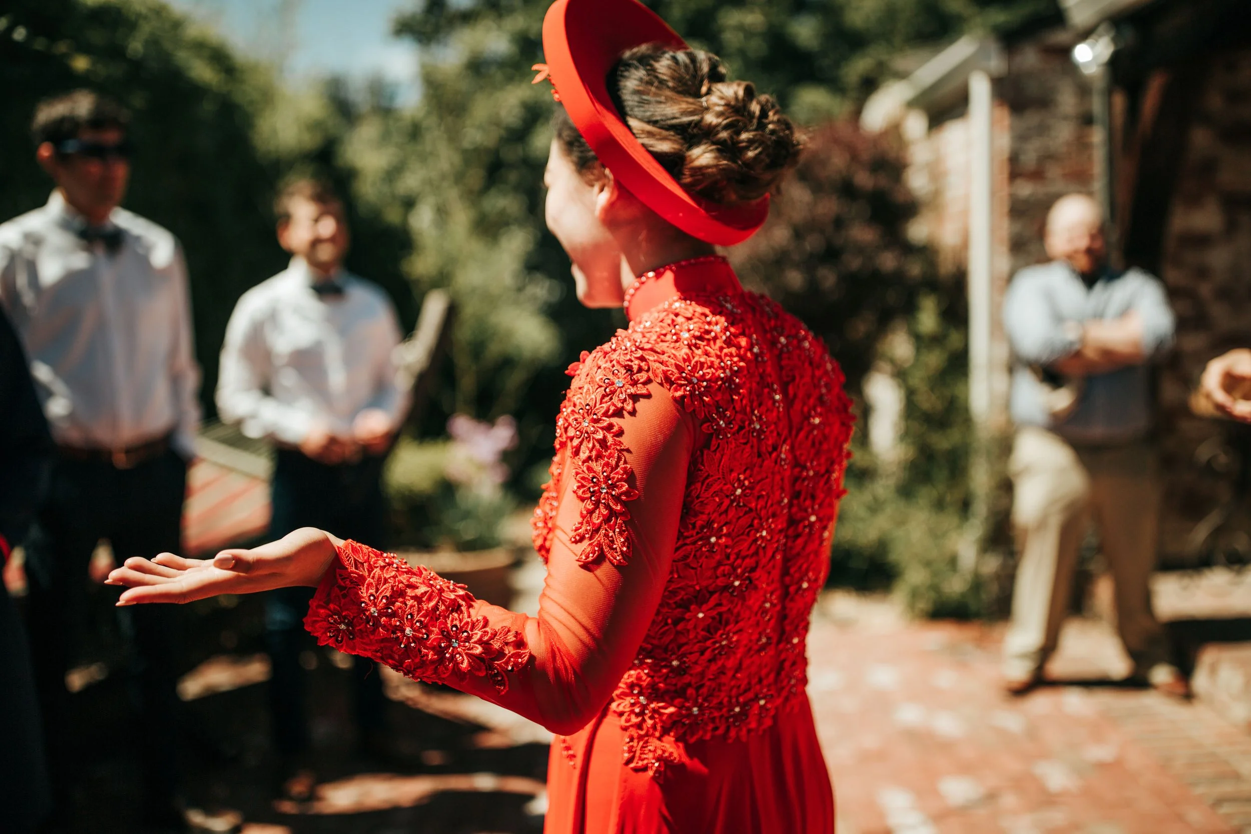 Woman in a red dress with floral embroidery, wearing a matching red hat, speaking to a group of people outdoors on a sunny day.