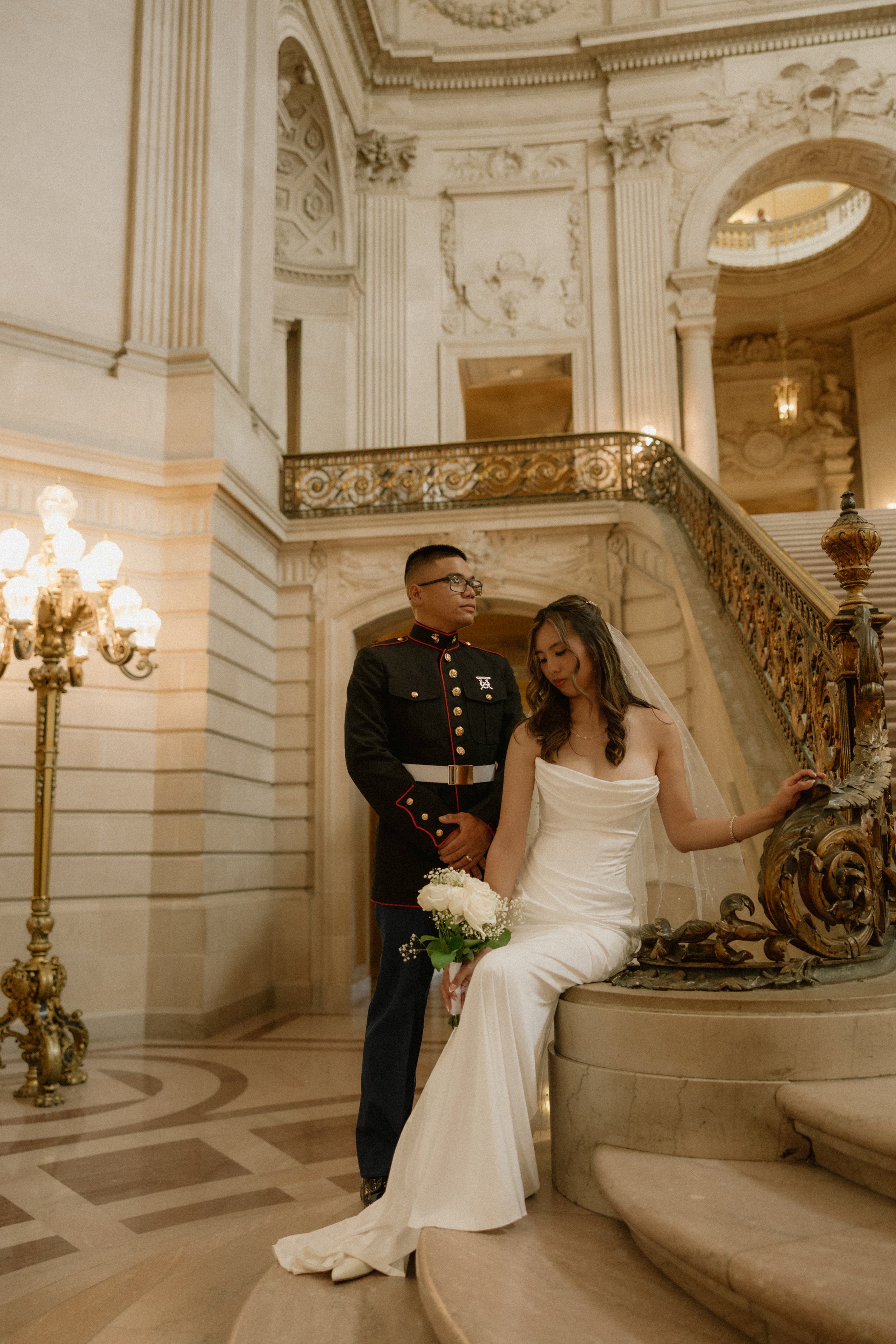 A couple standing on a grand staircase in an ornate, historic building. The man is in a military uniform, and the woman is in a white wedding gown holding a bouquet of white roses. Elegant gold and crystal chandelier lighting and intricate architectu