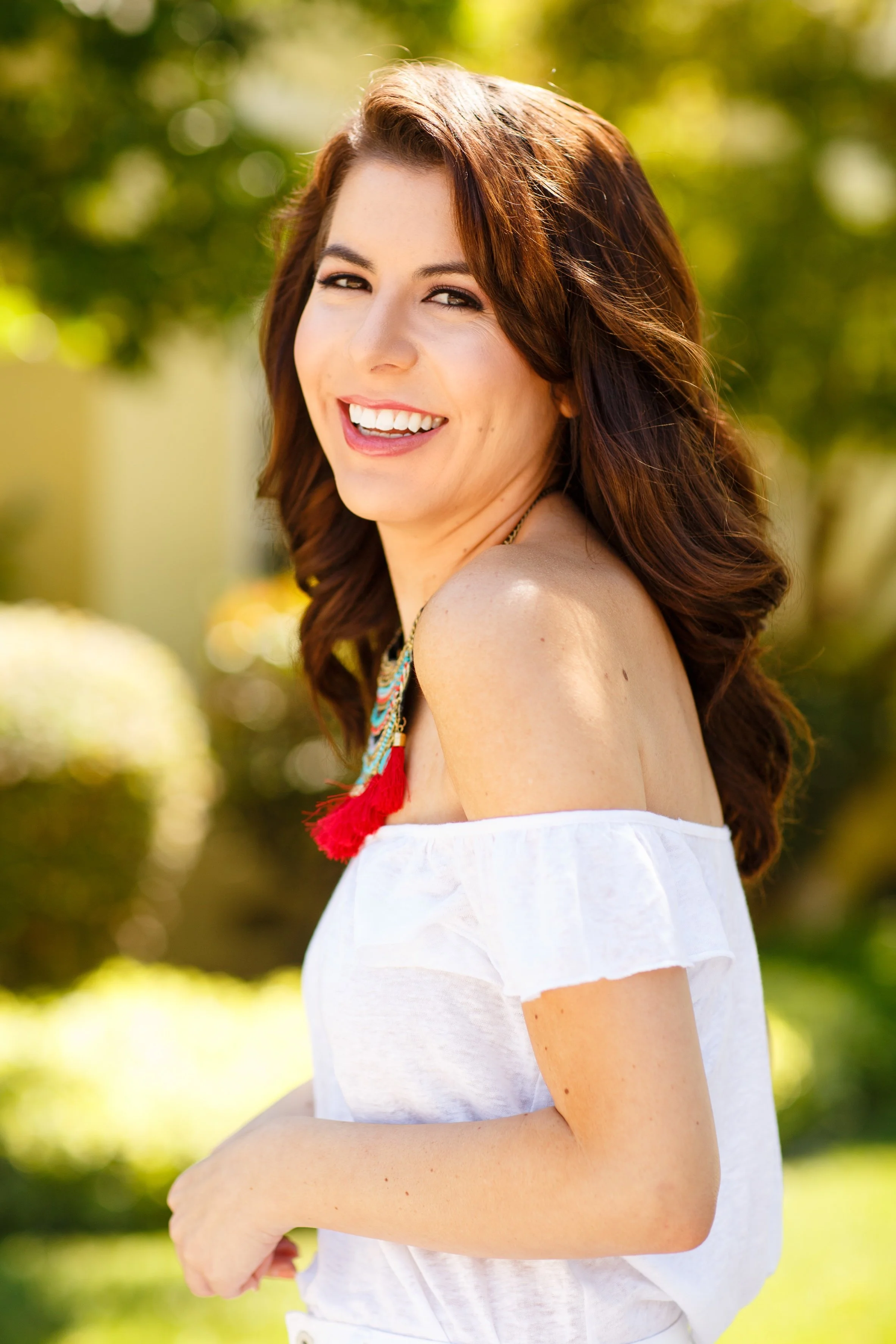 A young woman with long, wavy brown hair smiling outdoors in a sunny garden, wearing an off-shoulder white top and colorful necklace.