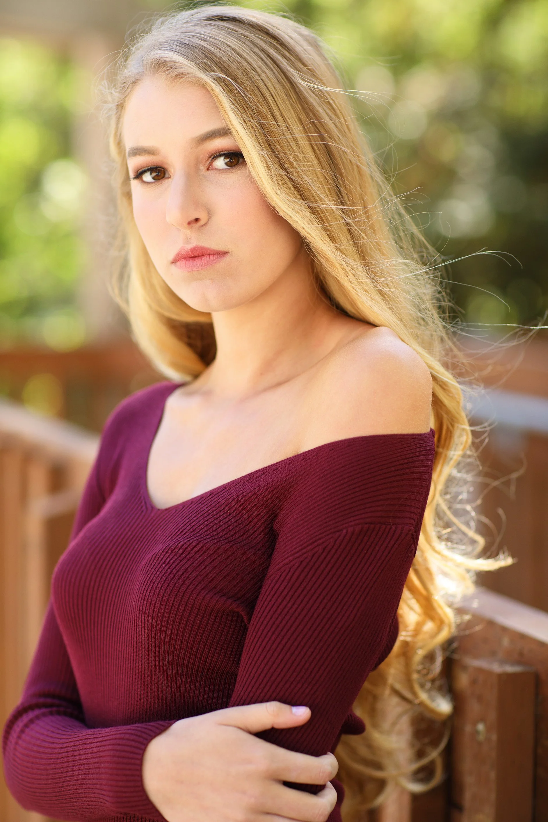 Young woman with long blonde hair wearing a burgundy off-the-shoulder sweater outdoors, standing near a wooden railing with greenery in the background.