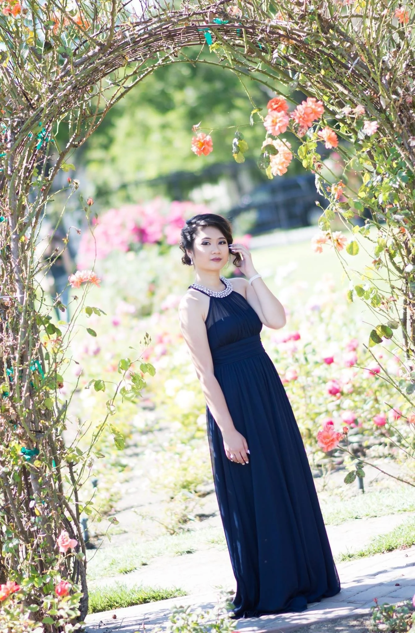 A woman in a navy blue gown standing under a flower arch in a garden with pink and peach roses.