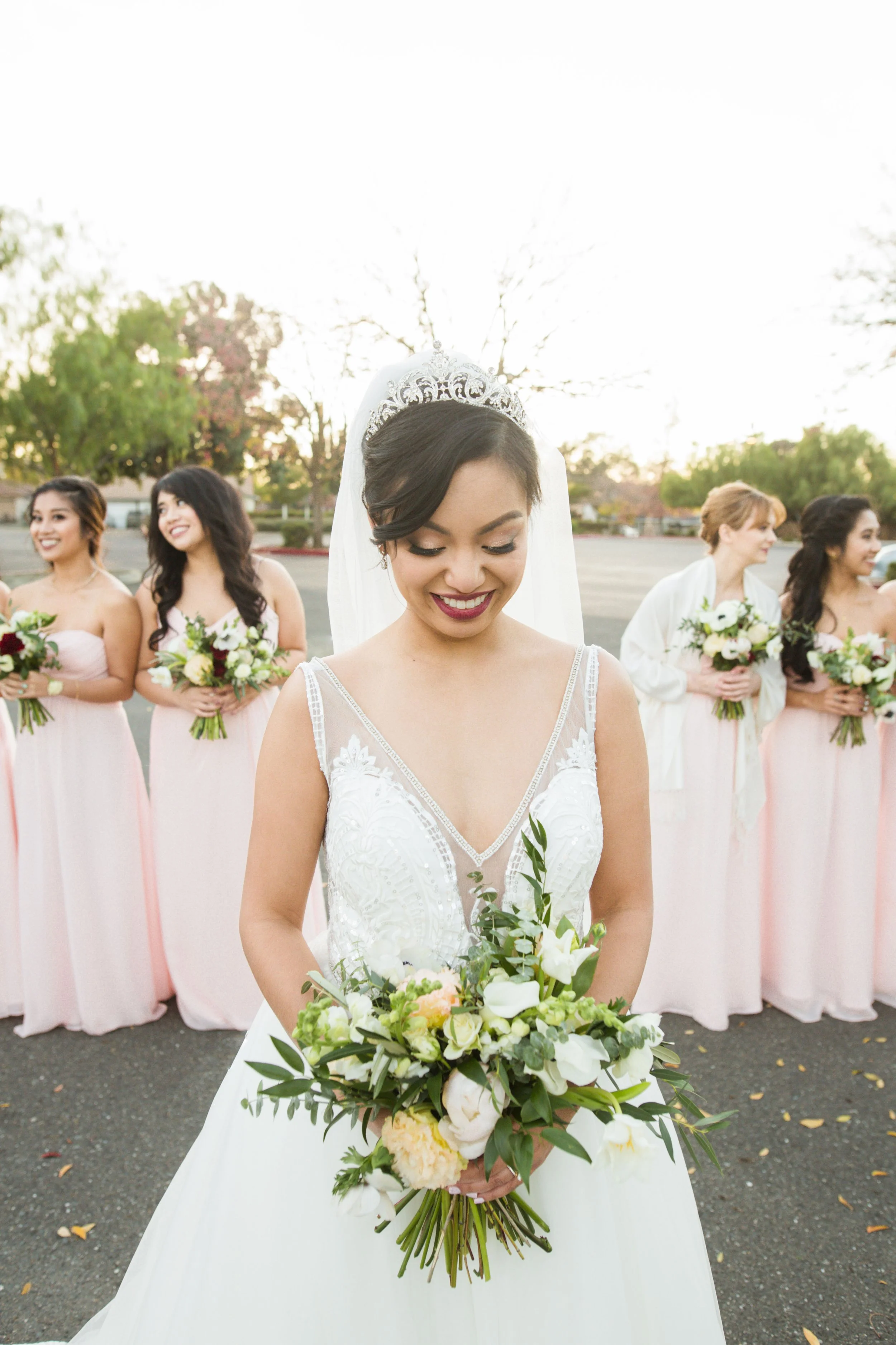 Bride in a white wedding dress holding a bouquet of white and peach flowers, surrounded by bridesmaids in pink dresses holding similar bouquets, outside during sunset.