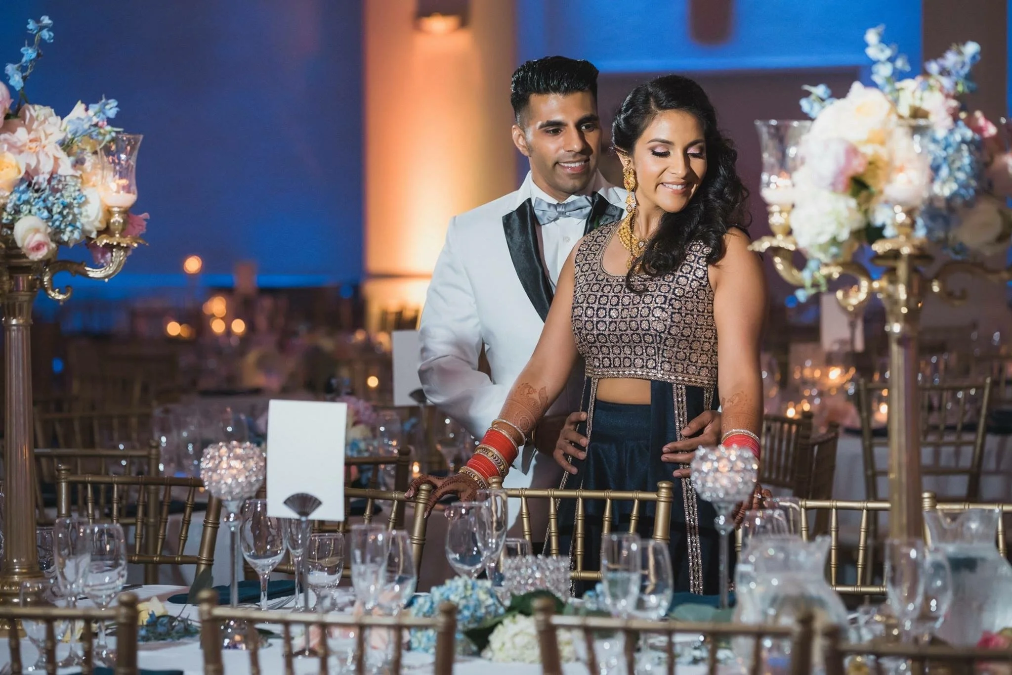 A couple dressed in formal attire standing at a decorated table during a celebration, with floral centerpieces and elegant tableware, in a warmly lit banquet hall.