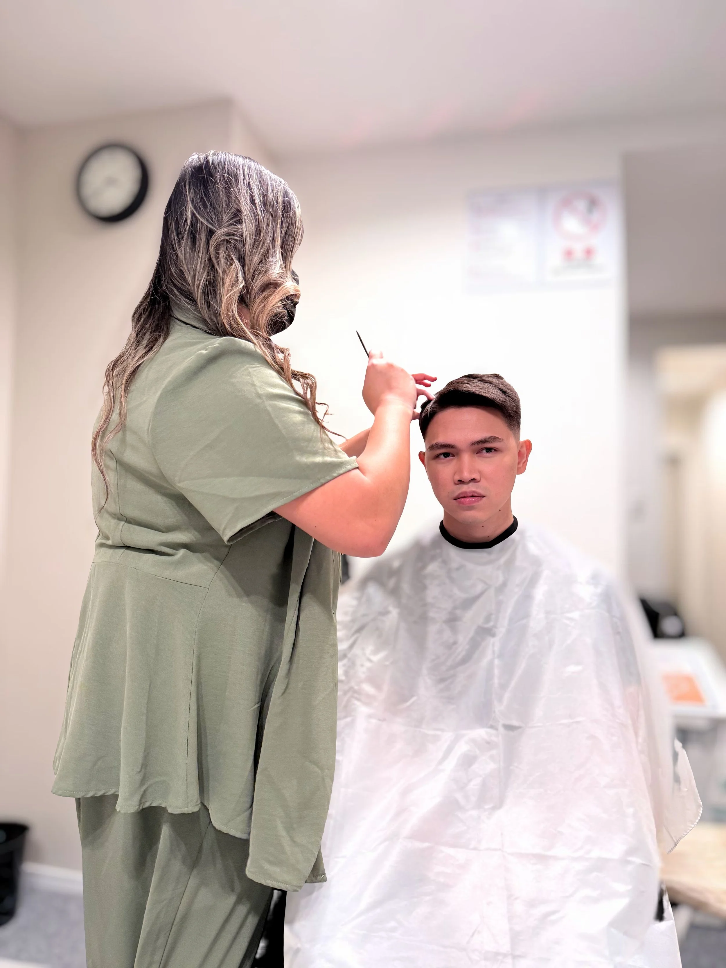A hairdresser cutting a young man's hair in a salon, with a clock on the wall in the background.