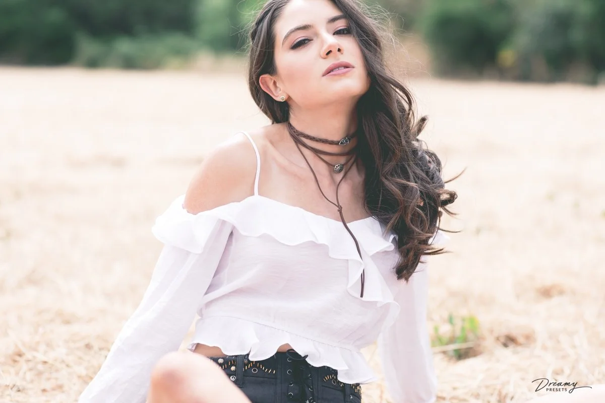 A young woman with long wavy brown hair posing outdoors on a sandy surface, wearing a white off-shoulder ruffled top, layered necklaces, and black shorts.