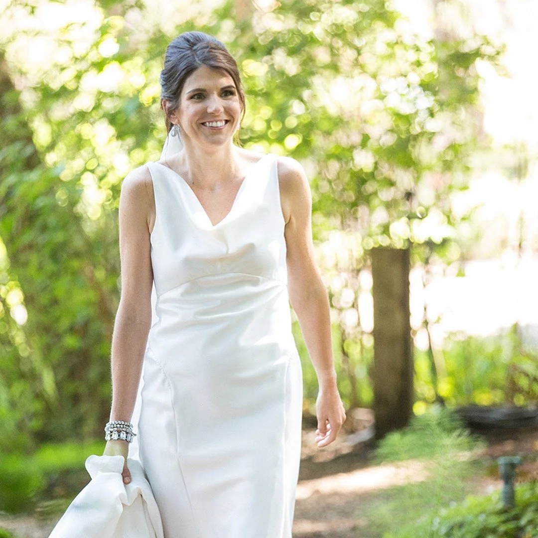 A woman in a white dress walking outdoors on a sunny day, surrounded by green trees and foliage, smiling and holding the hem of her dress.