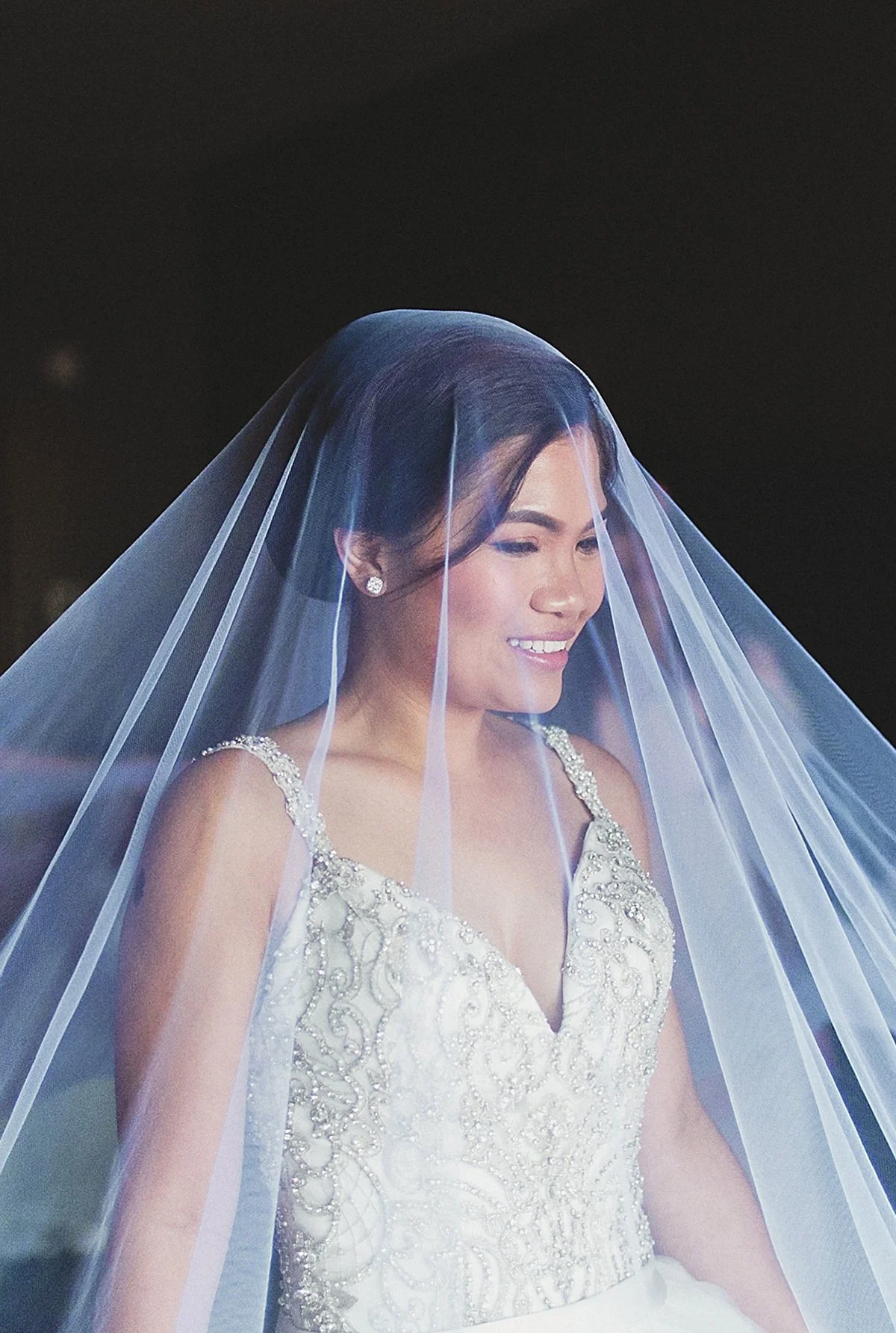 A woman in a wedding dress with a veil over her head, smiling gently.