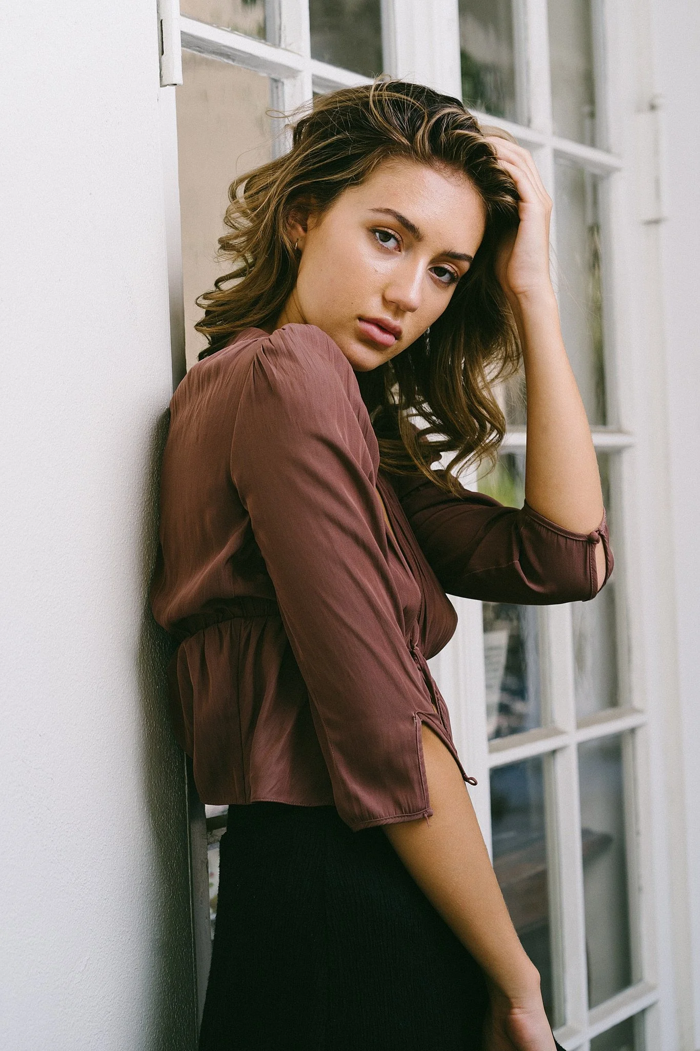 A young woman with wavy brown hair leaning against a white wall near a window, wearing a satin brown jacket and a black skirt.