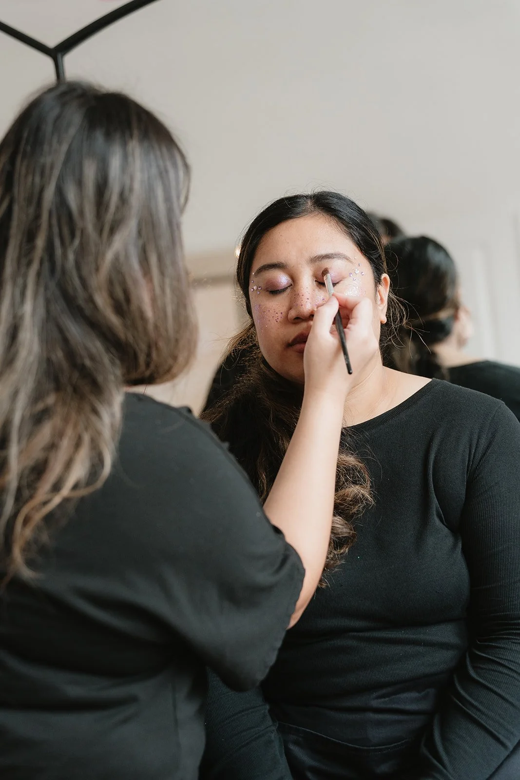 A woman with long dark hair is getting her makeup done by another woman with wavy hair, with glittery decorations around her eyes, wearing a black top.