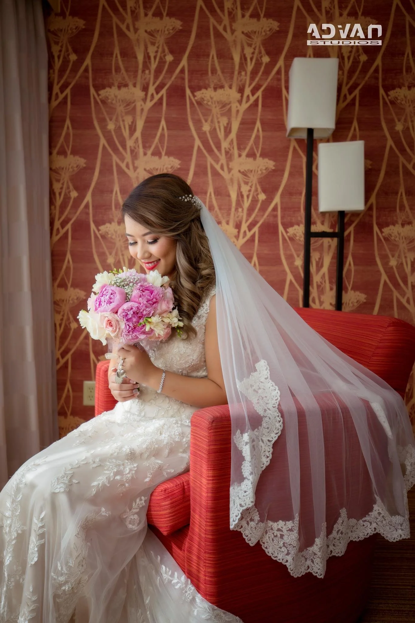 Bride sitting on a red armchair in a room with floral wallpaper, holding a bouquet of pink and white flowers, wearing a lace wedding dress and veil, smiling with her eyes closed.