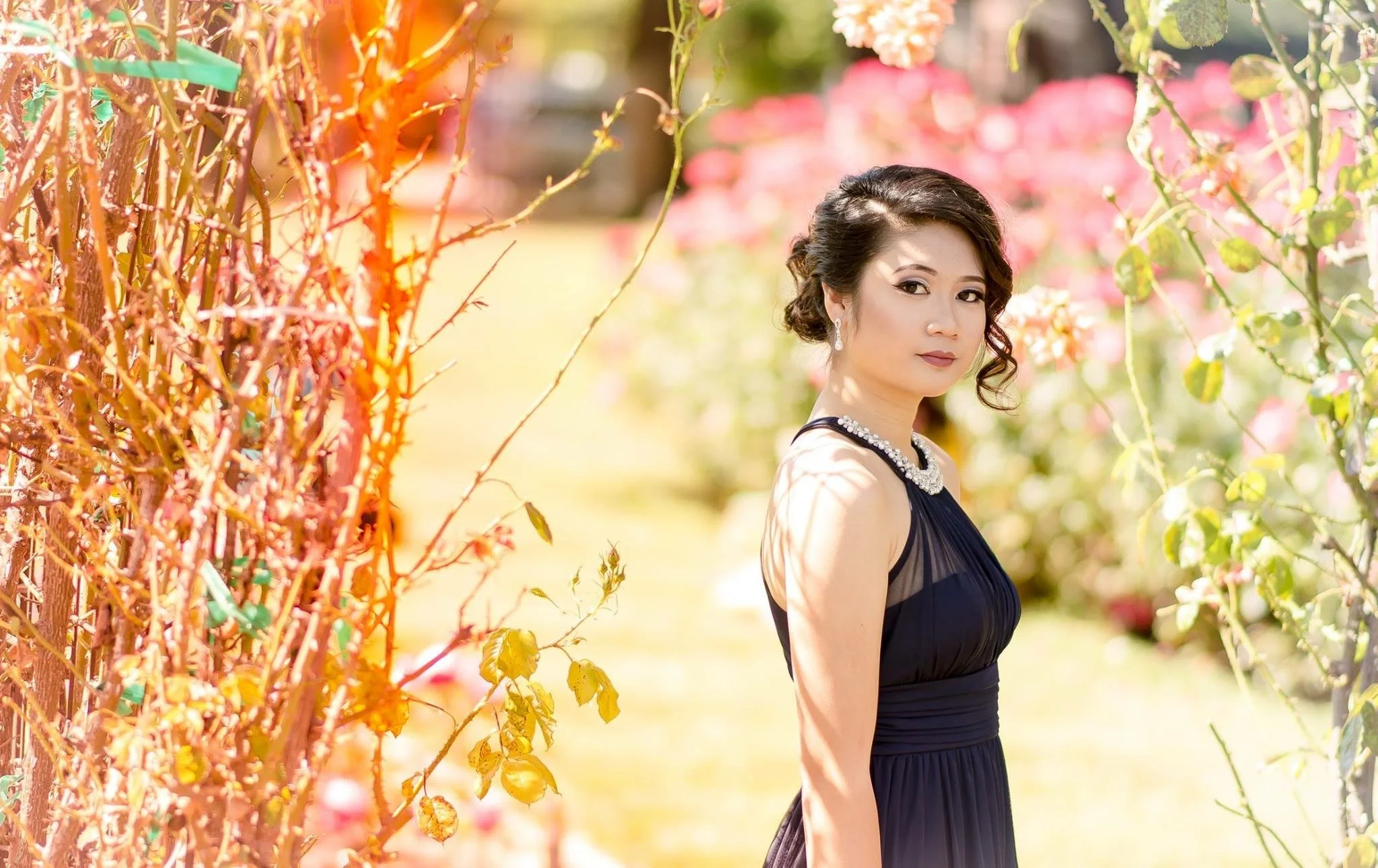 Woman in a black dress standing among pink and green flowering bushes in sunlight.