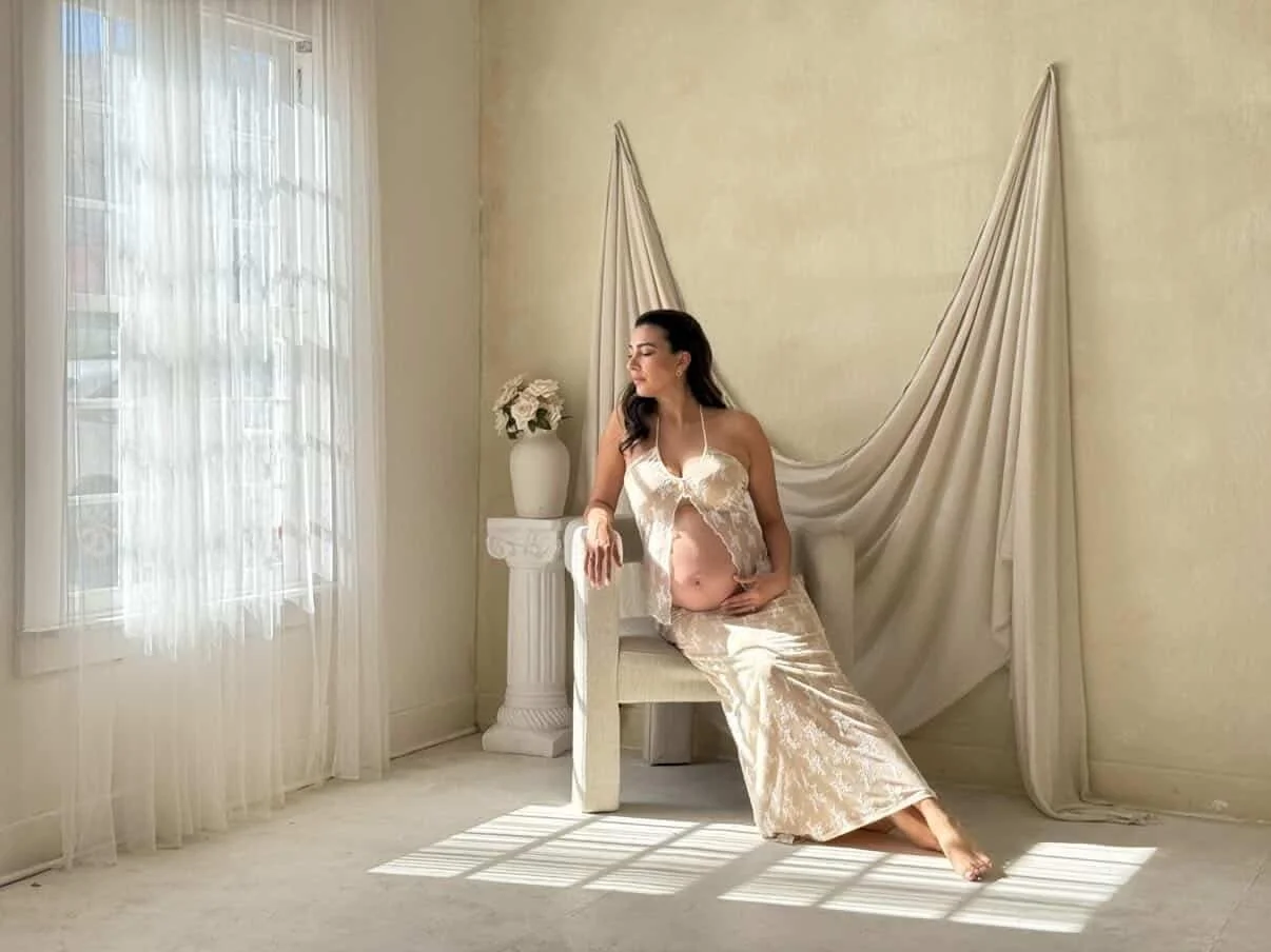 A woman in cream-colored satin and lace nightwear sitting on a chair, with her hand on her pregnant belly, in a softly lit room with sheer curtains and a large window.