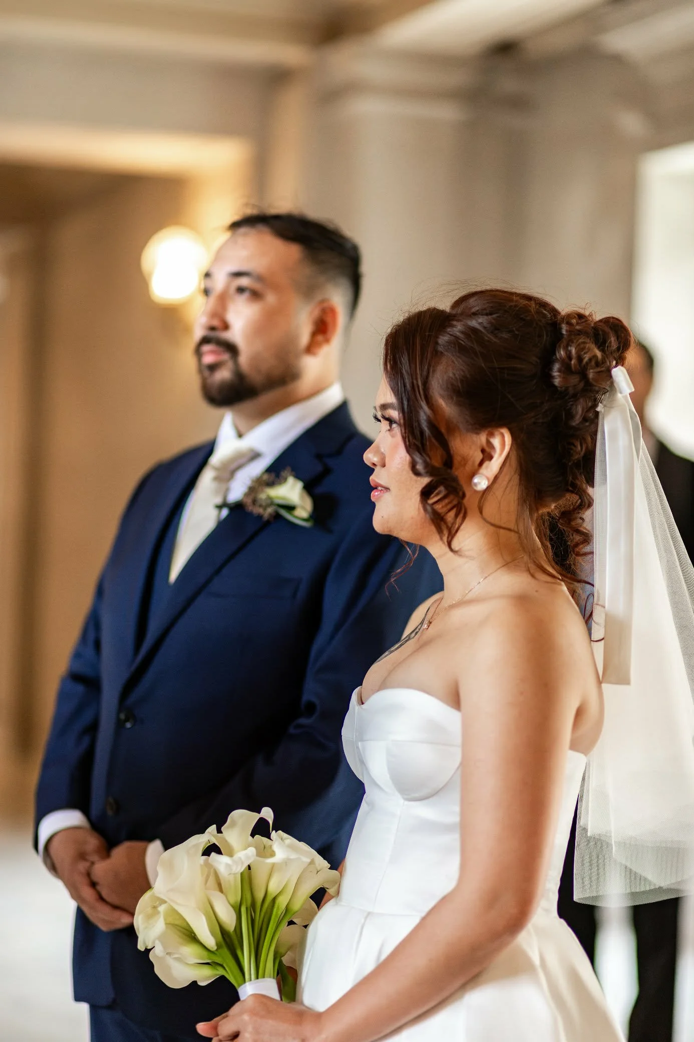 A bride and groom stand side by side during a wedding ceremony, with the bride holding a bouquet of white calla lilies. The groom is wearing a dark blue suit with a white shirt and a white tie, while the bride wears a strapless white wedding dress an