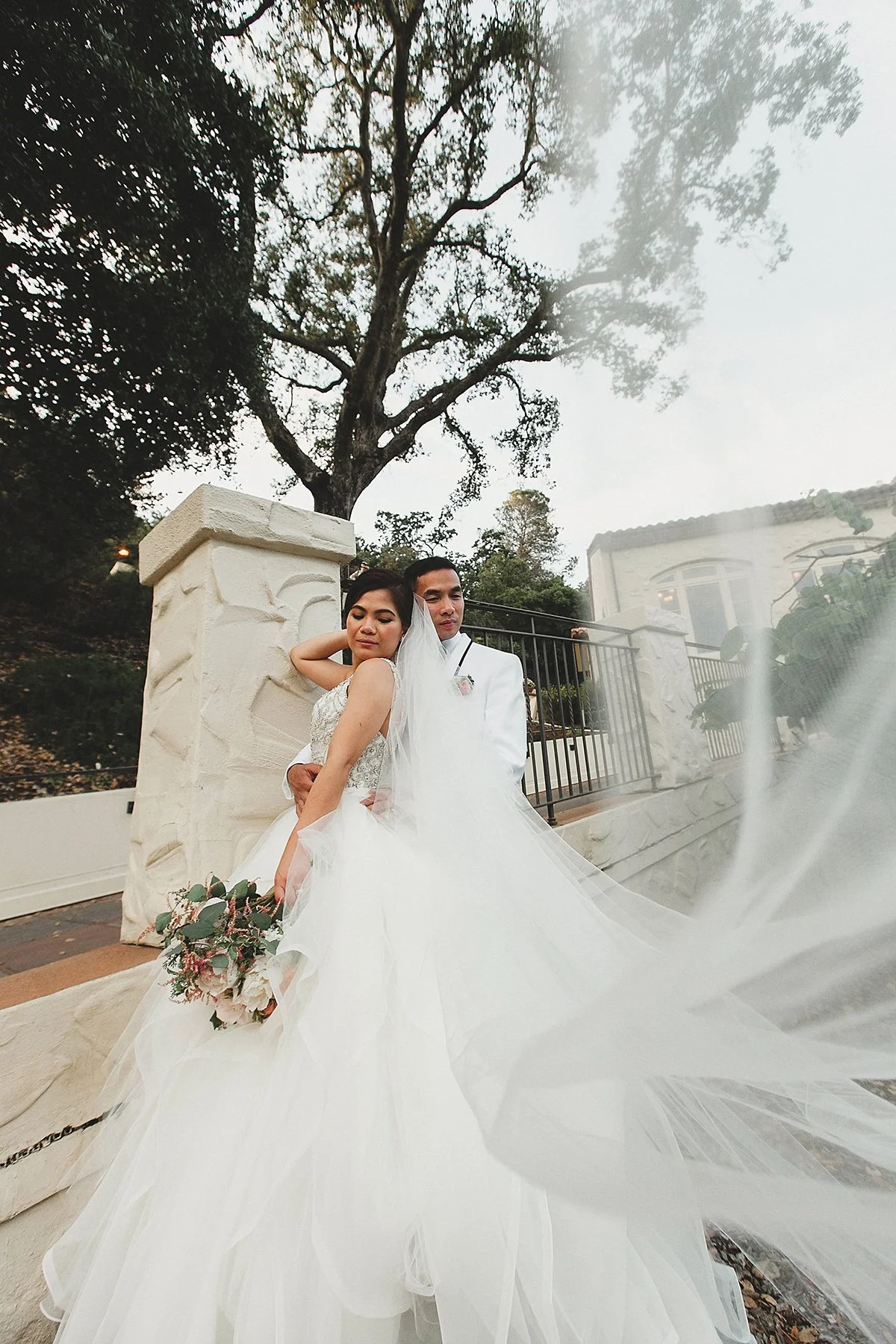 A bride and groom in wedding attire posing outdoors, with a large tree and a white building in the background.