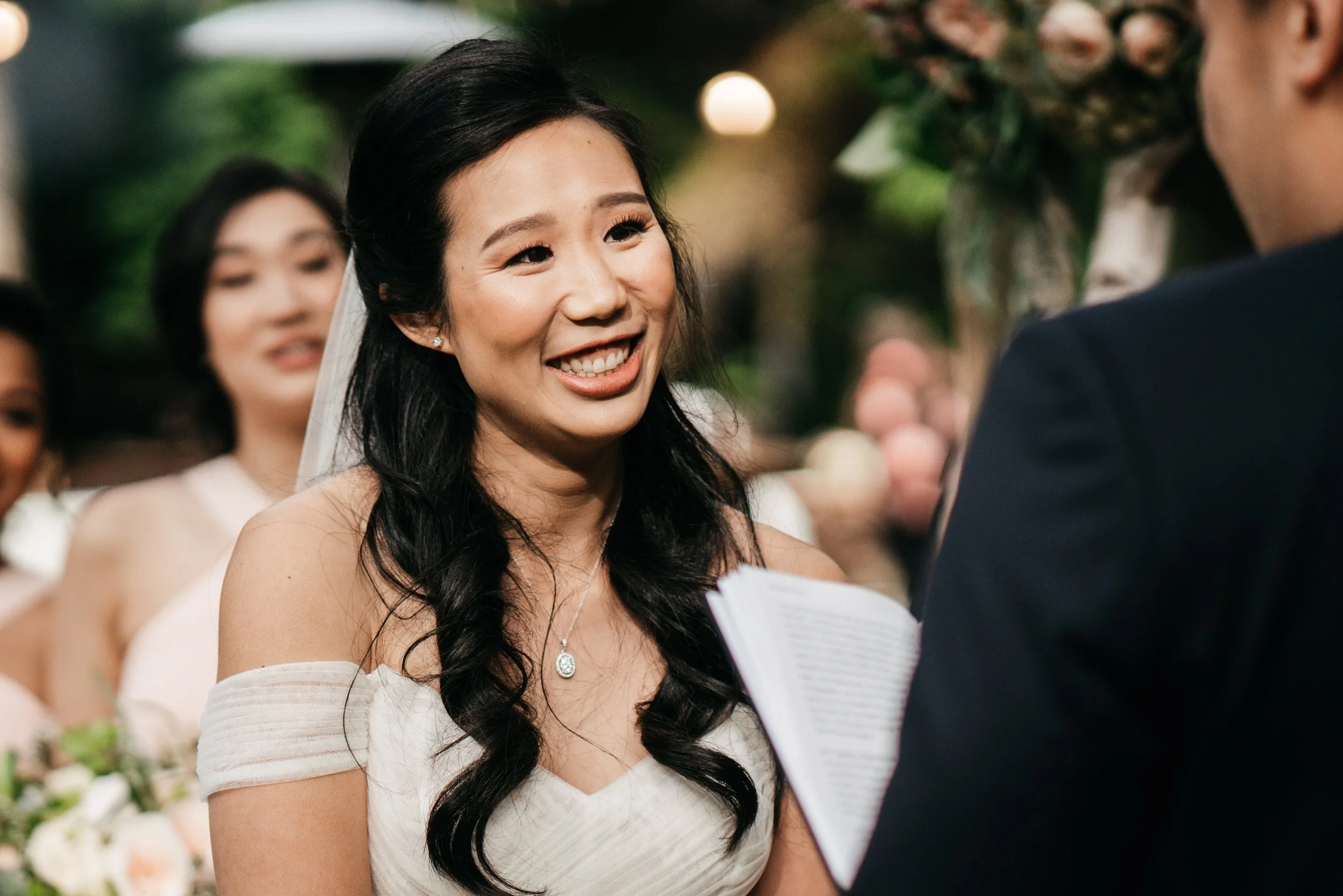 A bride smiling at the officiant during her wedding ceremony, with bridesmaids in the background.