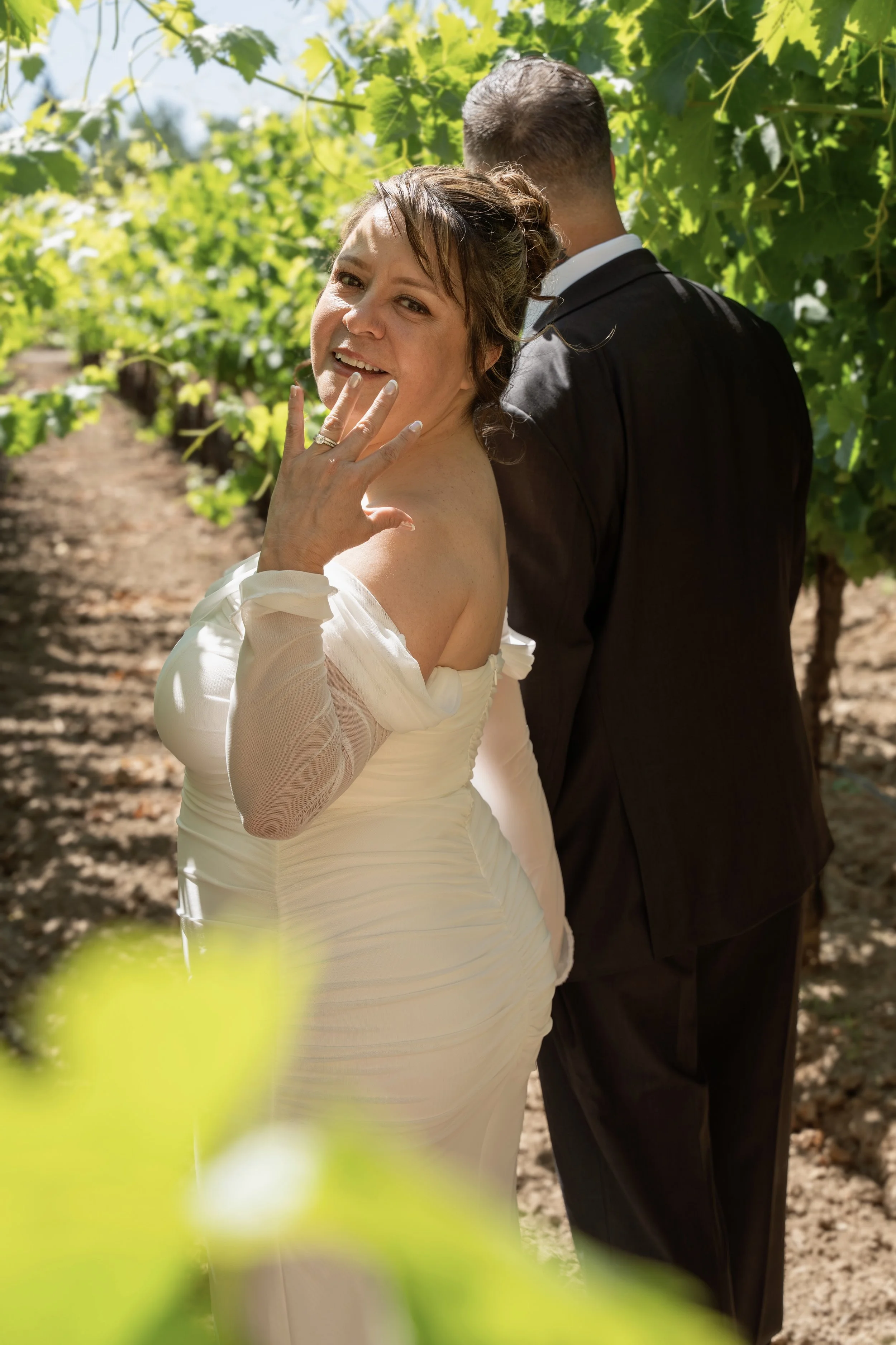 A woman in a wedding dress and a man in a suit stand back to back in a vineyard, with her smiling at the camera and her hand near her face, showing her wedding ring; the vineyard has green leaves and rows of grapevines.