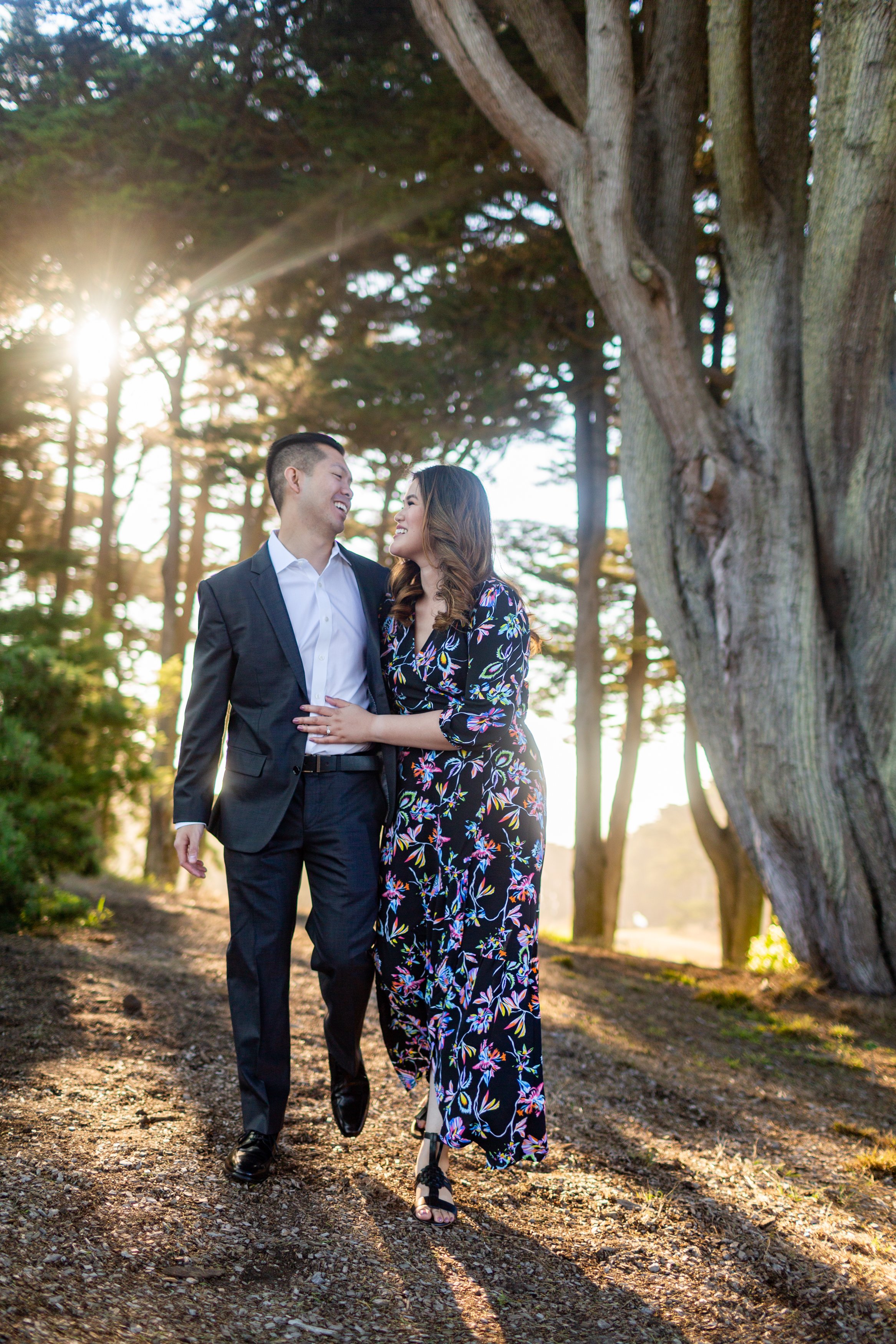 A couple walking outdoors together during sunset, smiling and looking at each other, with trees and sunlight in the background.