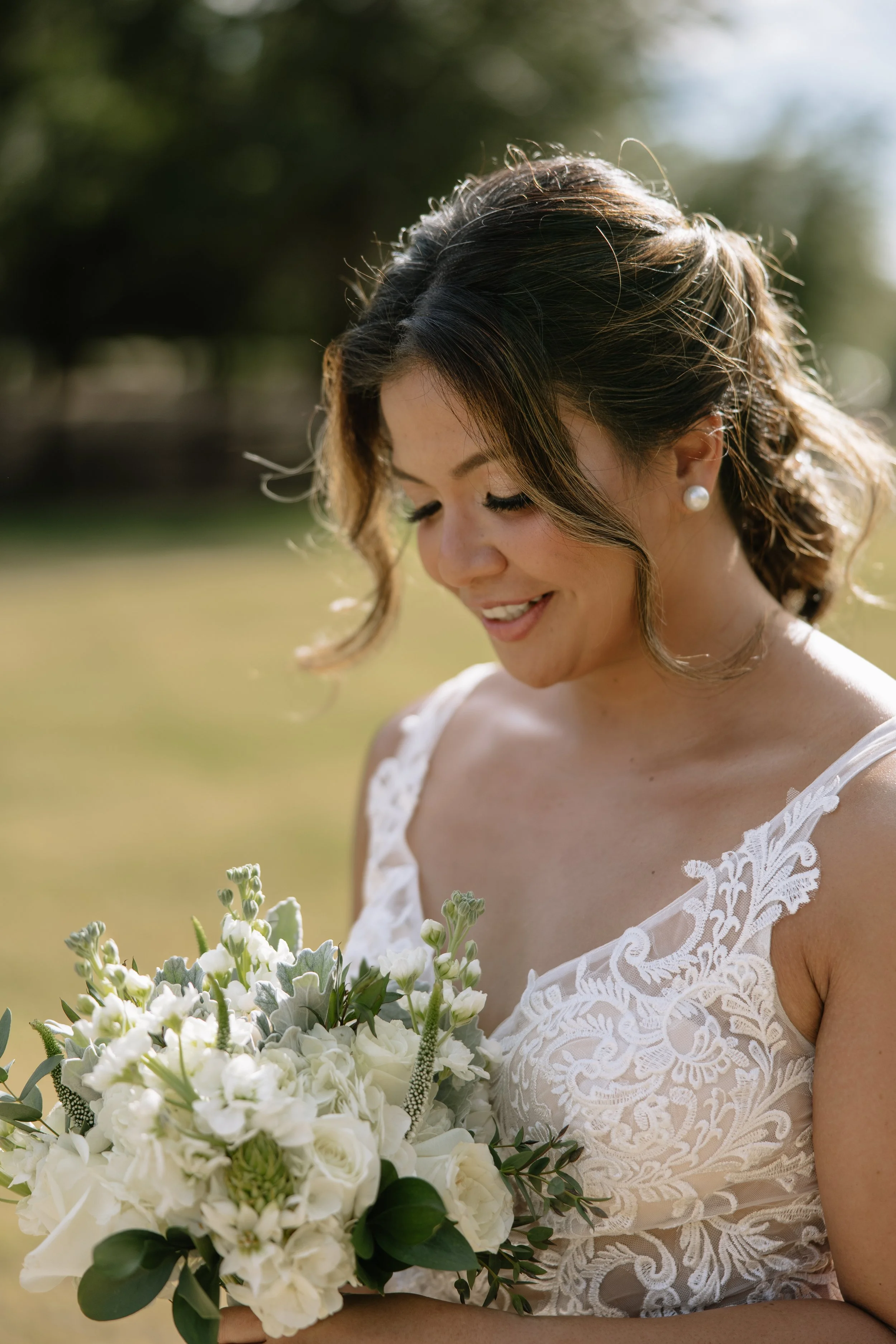 A bride holding a bouquet of white flowers, wearing a lace wedding dress, smiling and looking down outdoors in sunlight.