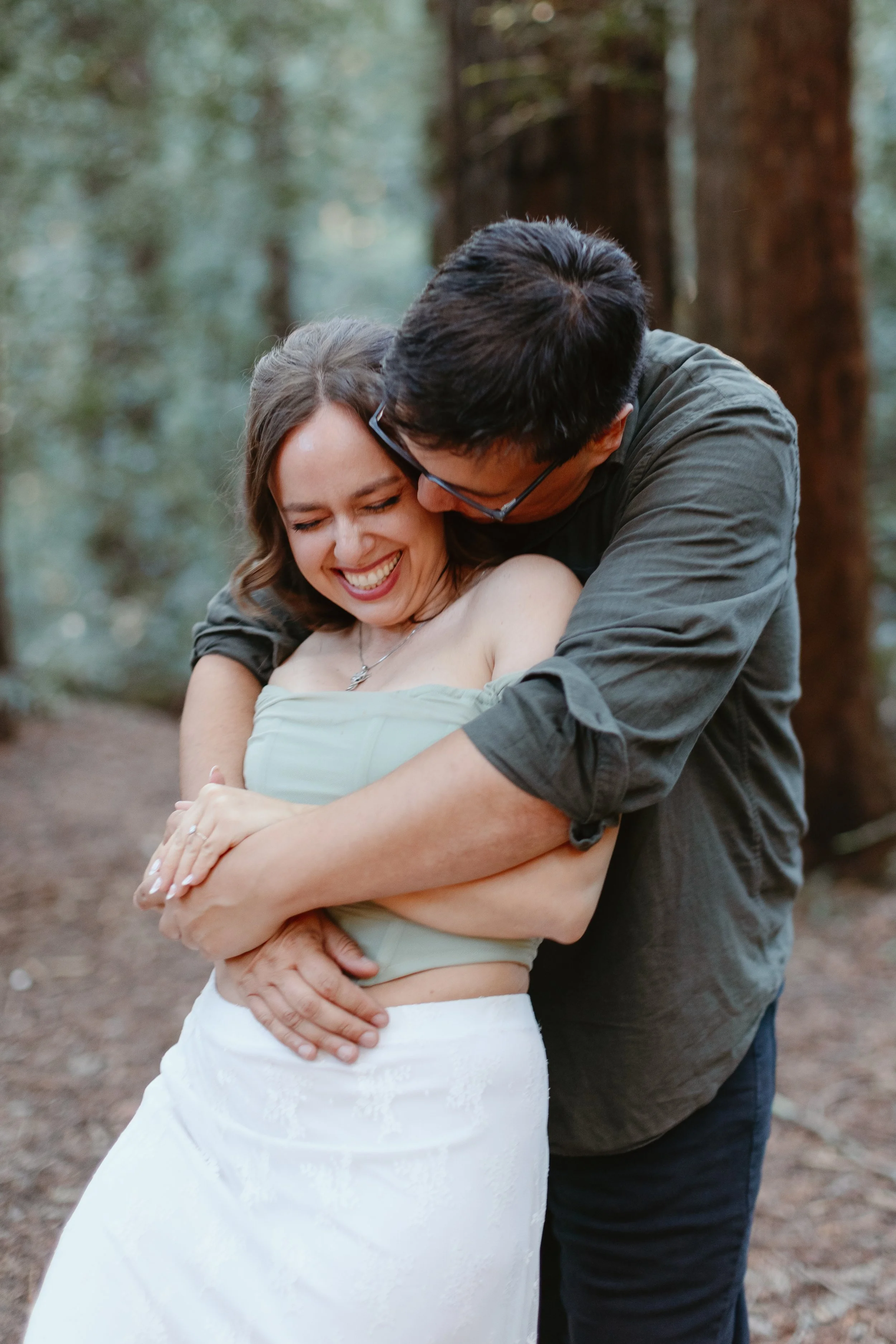 A man and a woman hugging outdoors in a forest, both smiling with eyes closed. The woman is wearing a strapless top and a white skirt, and the man is wearing glasses and a gray shirt.