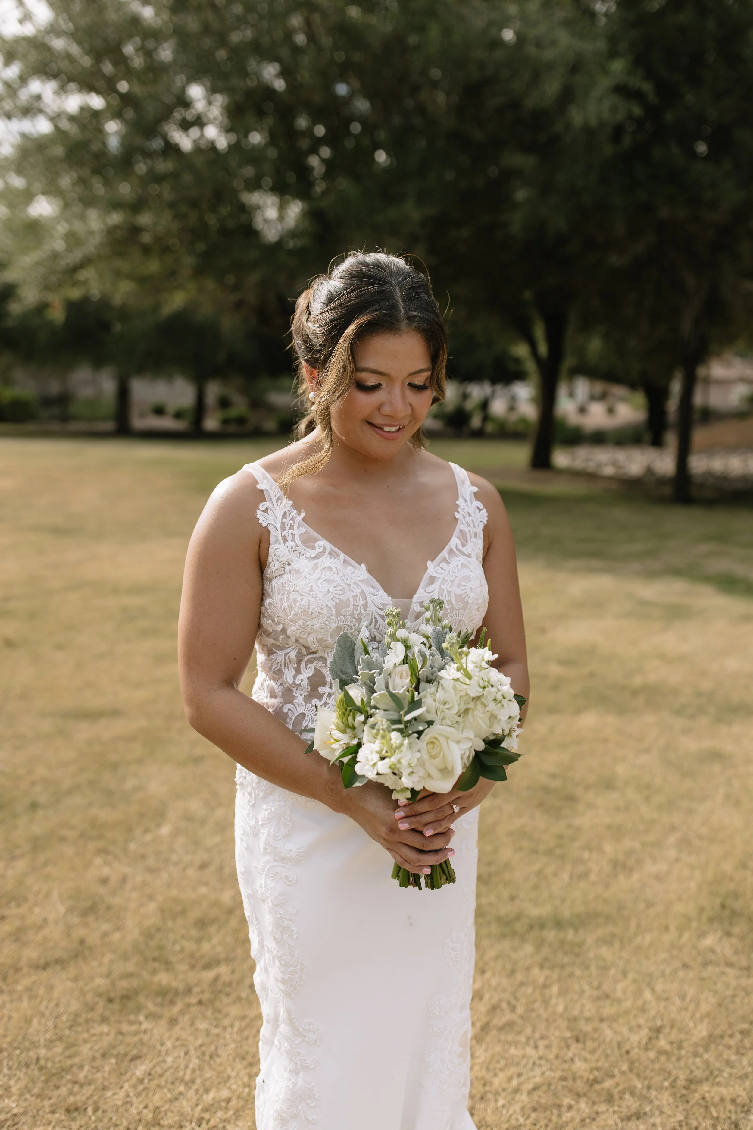 A bride in a white lace wedding dress holding a bouquet of white flowers standing on grass with trees in the background.