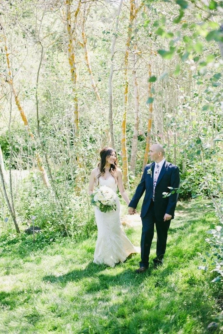 A bride and groom holding hands and smiling at each other in a lush outdoor setting with green grass and trees.