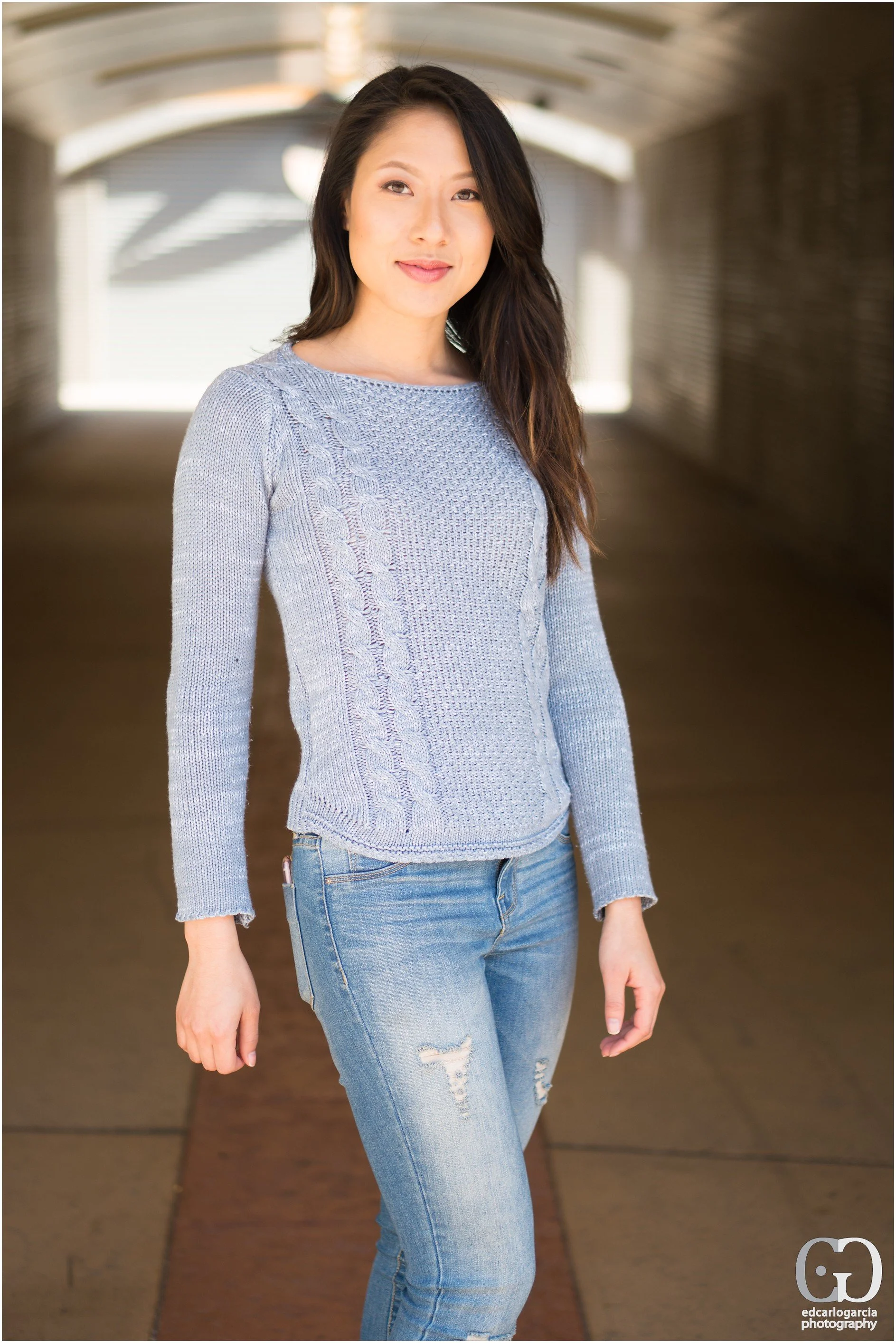 A young woman with long dark hair and light skin standing in a covered walkway, smiling at the camera, wearing a light blue sweater and ripped jeans.