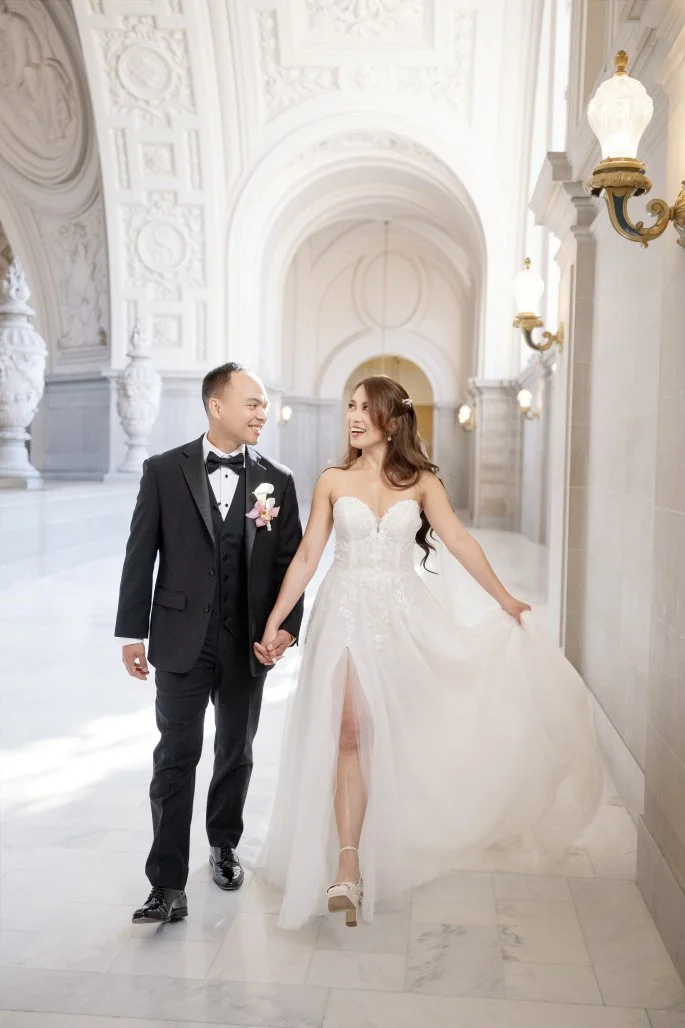 A bride and groom walking hand in hand in a bright, ornate hall. The bride wears a strapless white wedding gown with a slit, and the groom wears a black tuxedo with a bow tie. They are smiling at each other.