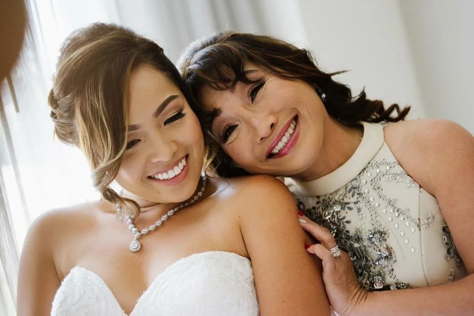 A young woman in a white wedding dress and a pearl necklace with an older woman in a white dress with silver embellishments, both smiling and leaning their heads together.