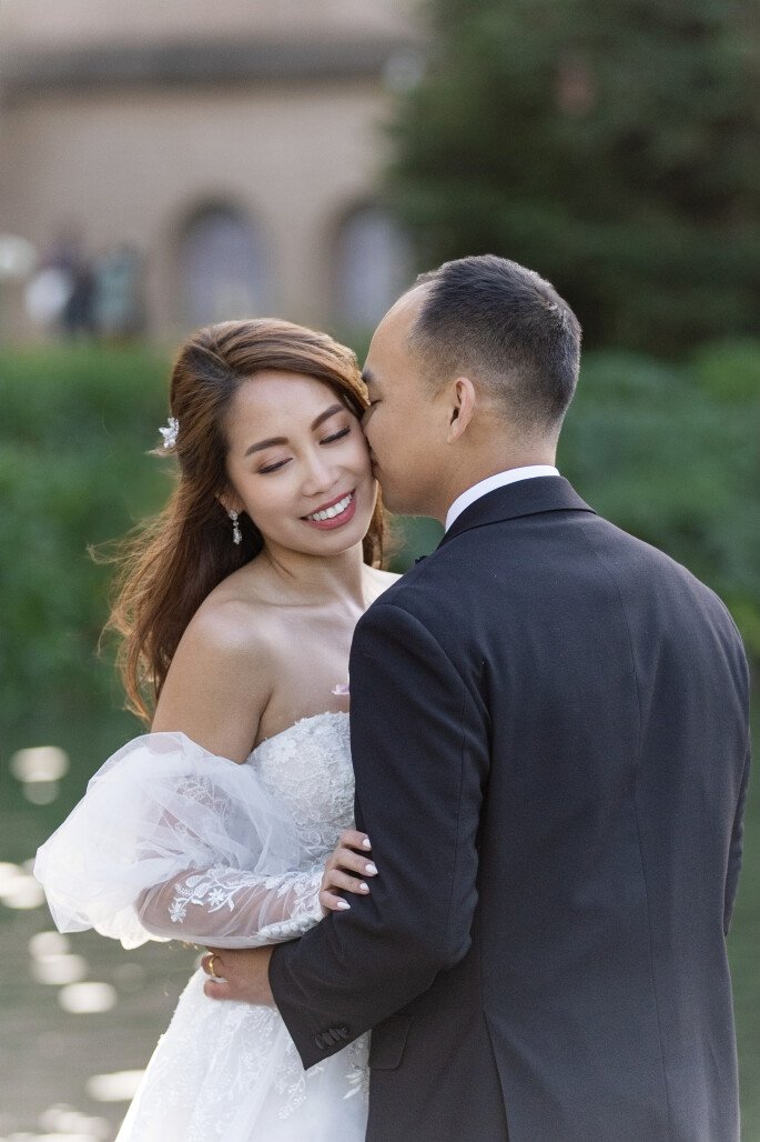 A couple in wedding attire sharing an intimate moment outdoors, with the groom kissing the bride's cheek and the bride smiling with her eyes closed.