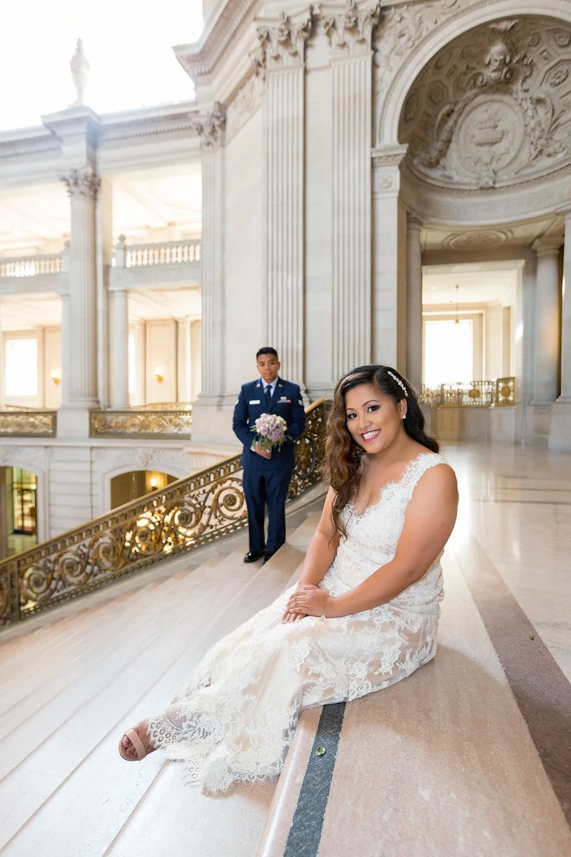 A woman in a wedding dress sitting on a marble ledge inside a grand, historic building with classical architecture, while a man in a military uniform holding a bouquet stands behind her.