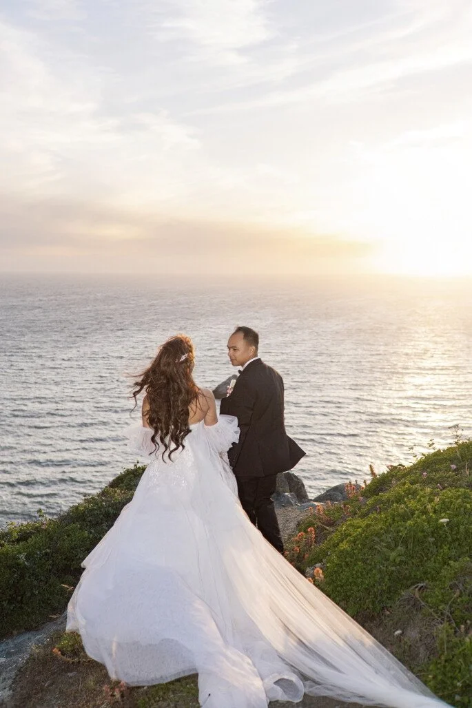 A bride and groom standing on a grassy cliff overlooking the ocean at sunset, holding hands and looking at each other.