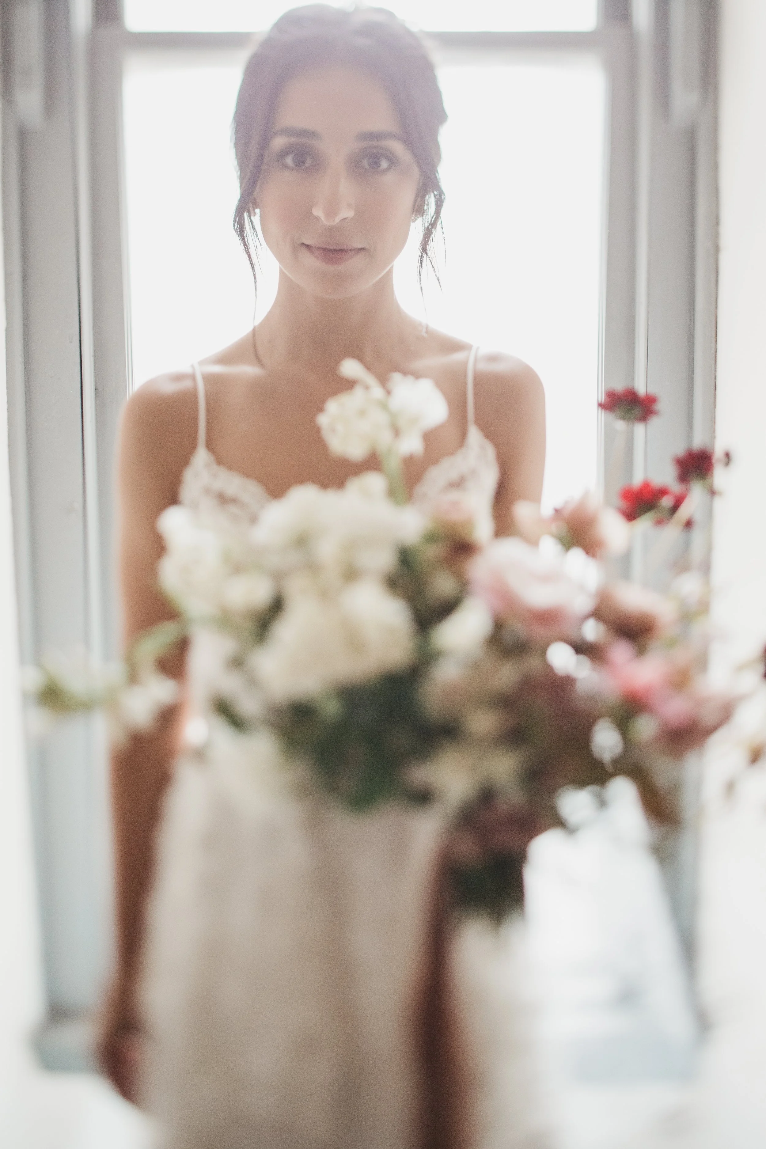 A woman in a white dress with lace straps looking at the camera while holding a bouquet of white and pink flowers, standing in front of a window.