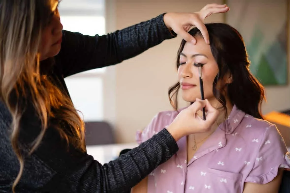 A woman getting her makeup done by a makeup artist in a indoor setting.