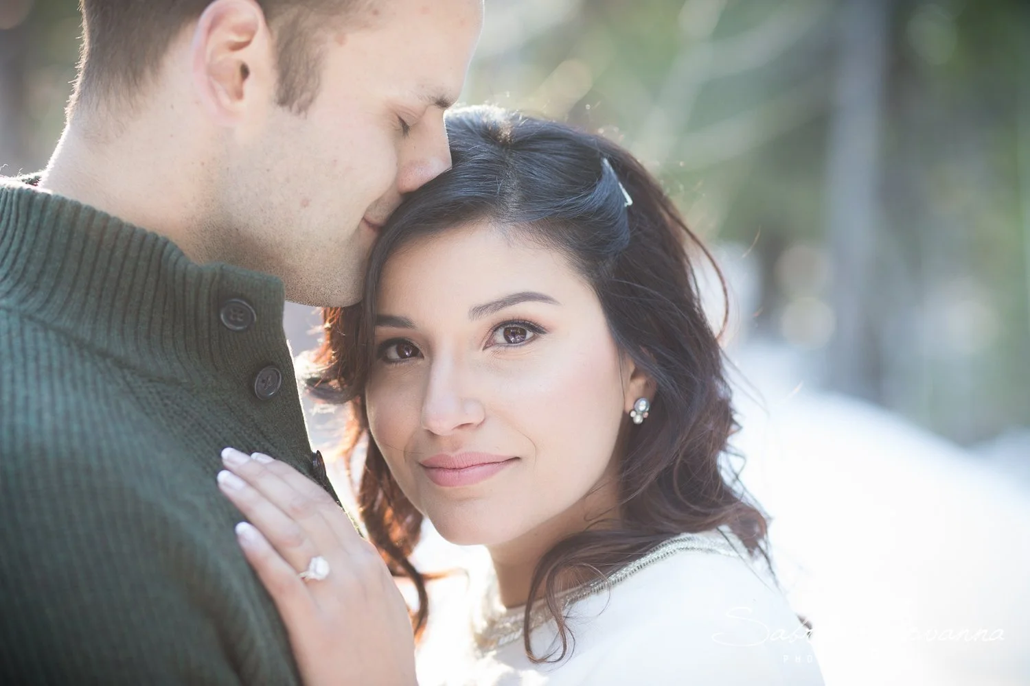 A couple embracing outdoors in winter, with the man gently resting his forehead on the woman's head, and the woman looking at the camera with a slight smile, sunlight shining through trees in the background.