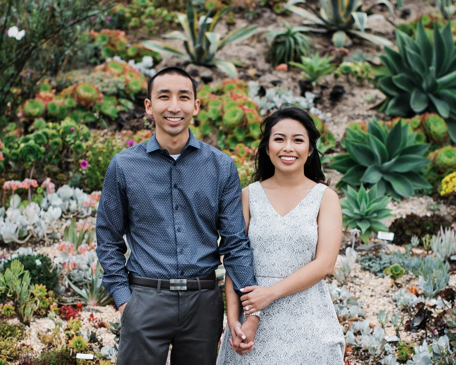 A smiling man and woman standing hand-in-hand in a garden filled with various succulents and cacti.