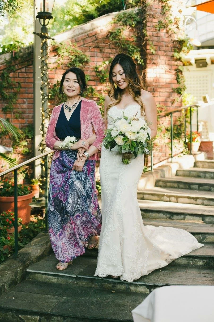 A bride in a white wedding dress holding a bouquet, walking down outdoor stone steps with an older woman in colorful attire, both smiling. They are in a garden setting with brick walls, plants, and fairy lights.